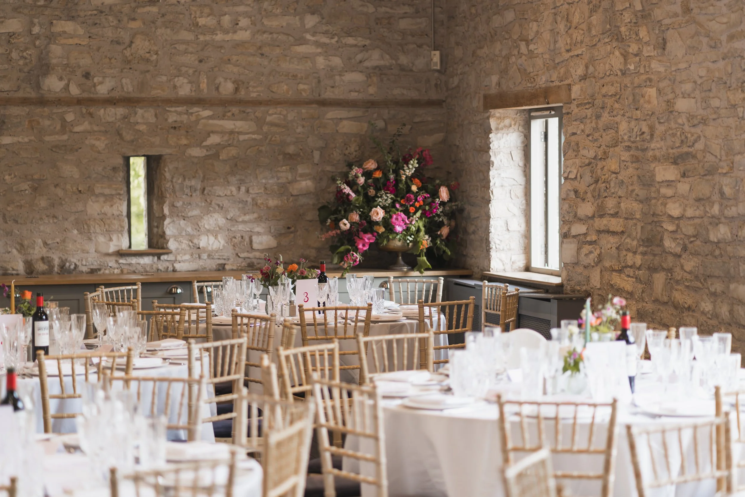 Interior of a rustic event space with exposed stone walls, large flower arrangements, and banquet tables set for a formal event with white tablecloths, glassware, and bottles of wine.