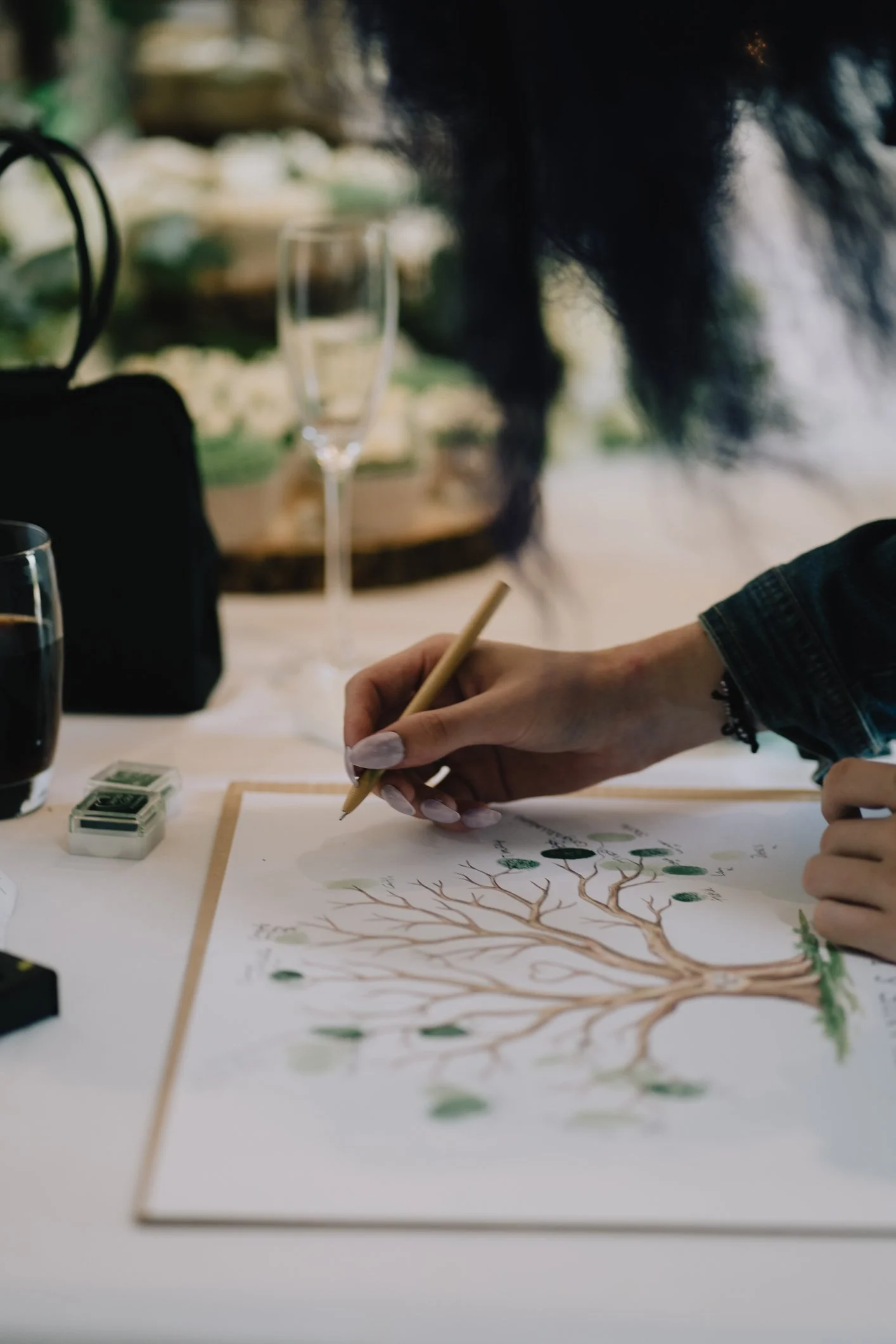 A person is drawing a tree with green leaves on a sheet of paper using a pencil.