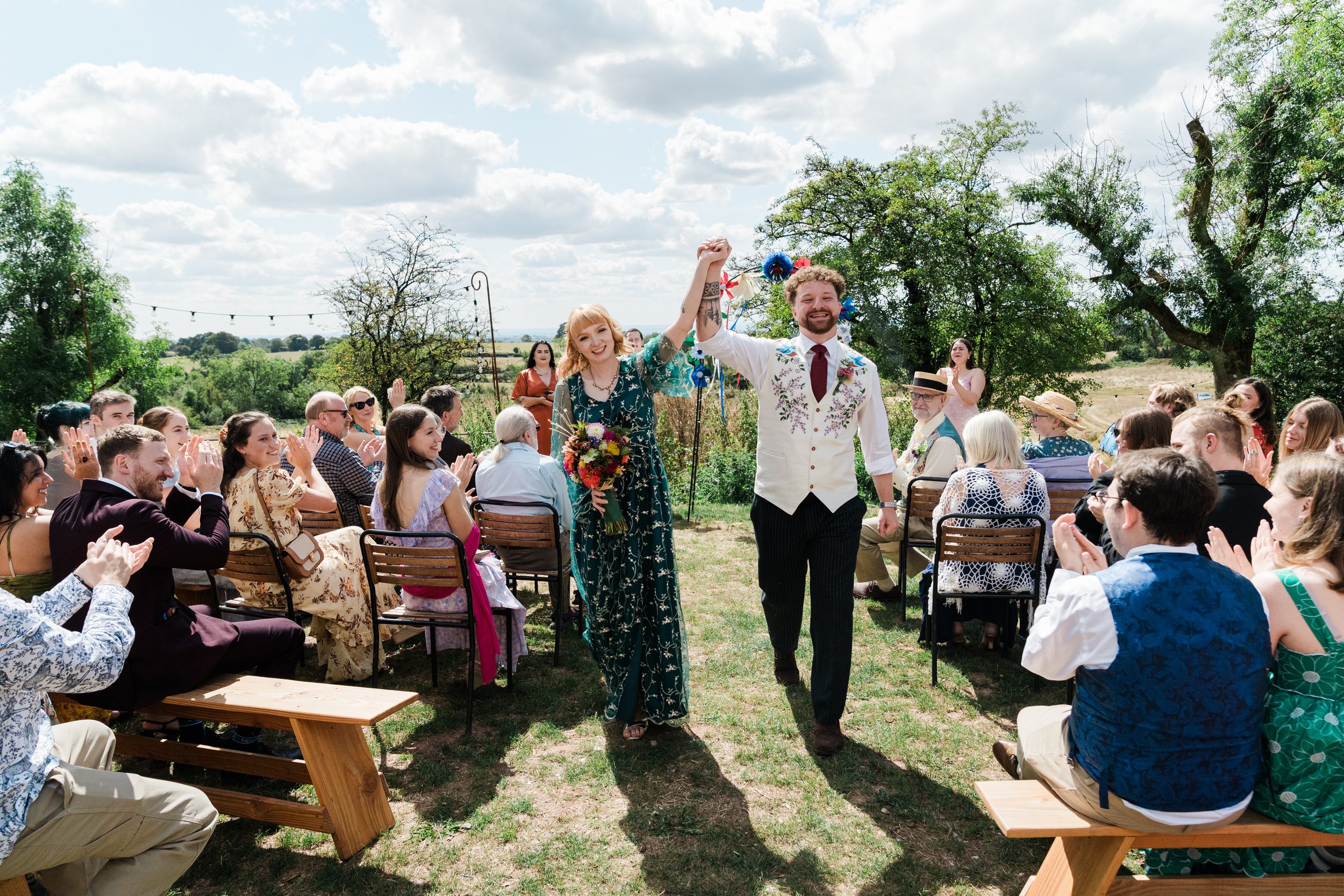 A wedding ceremony outdoors with the bride and groom holding hands and smiling, surrounded by seated guests clapping and celebrating under a partly cloudy sky, with trees and a scenic landscape in the background.
