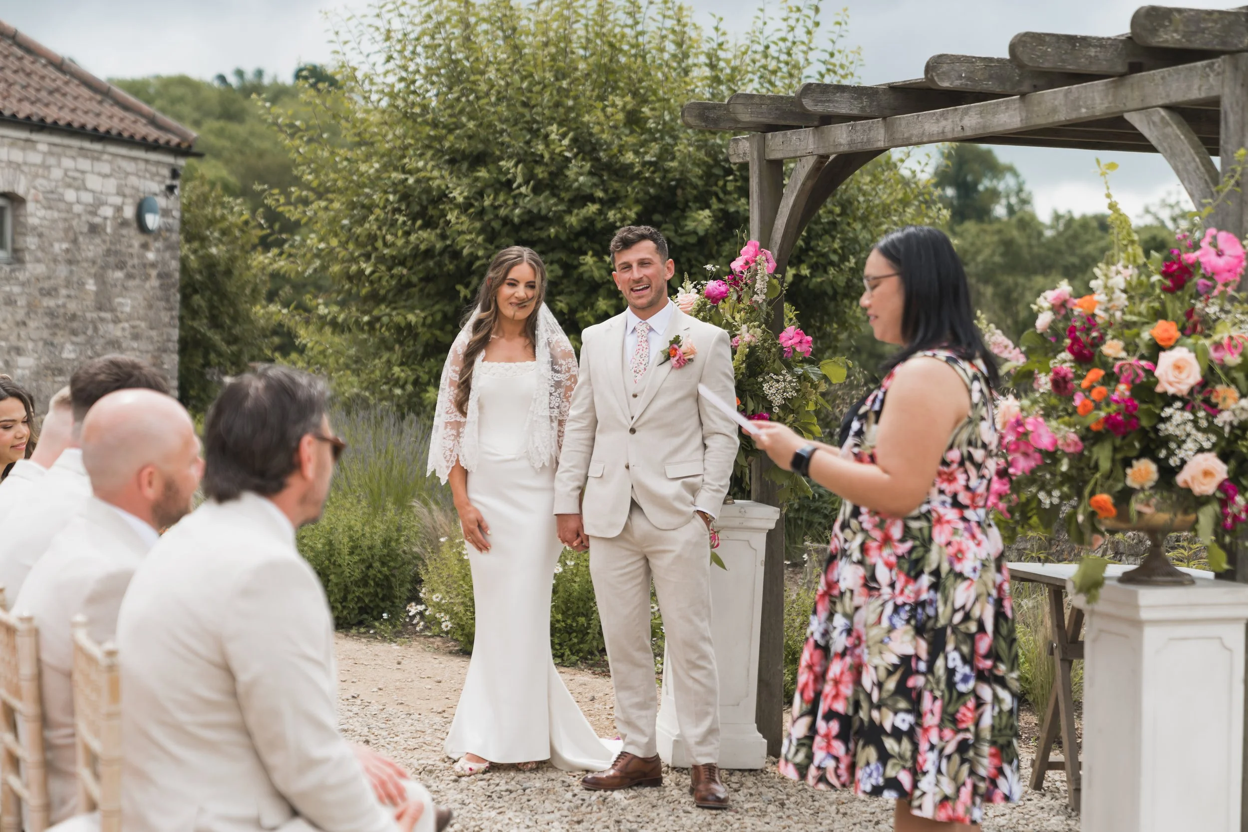 A wedding ceremony outdoors with a bride and groom holding hands, standing under a wooden arch decorated with pink and white flowers, facing an officiant reading from a paper, with guests seated nearby.