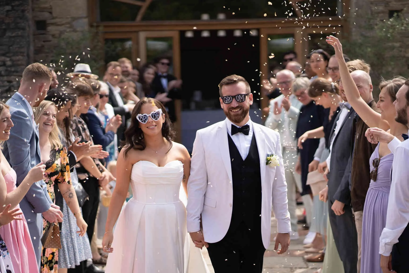 A bride and groom walking hand in hand through a crowd of guests, celebrating their wedding, as confetti falls around them at Oxleaze Barn.