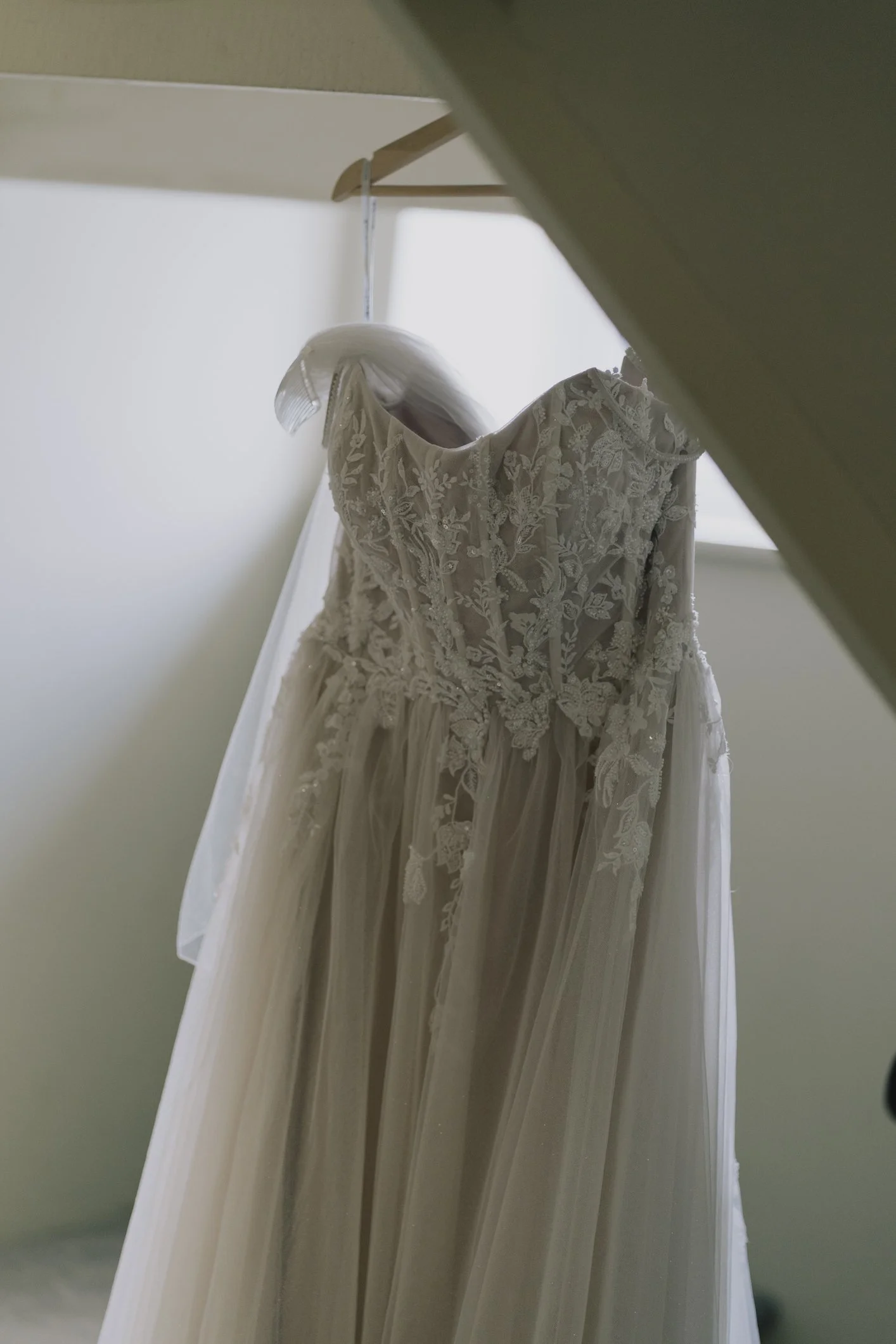 A lace wedding dress hanging on a wooden hanger against a light-colored wall.