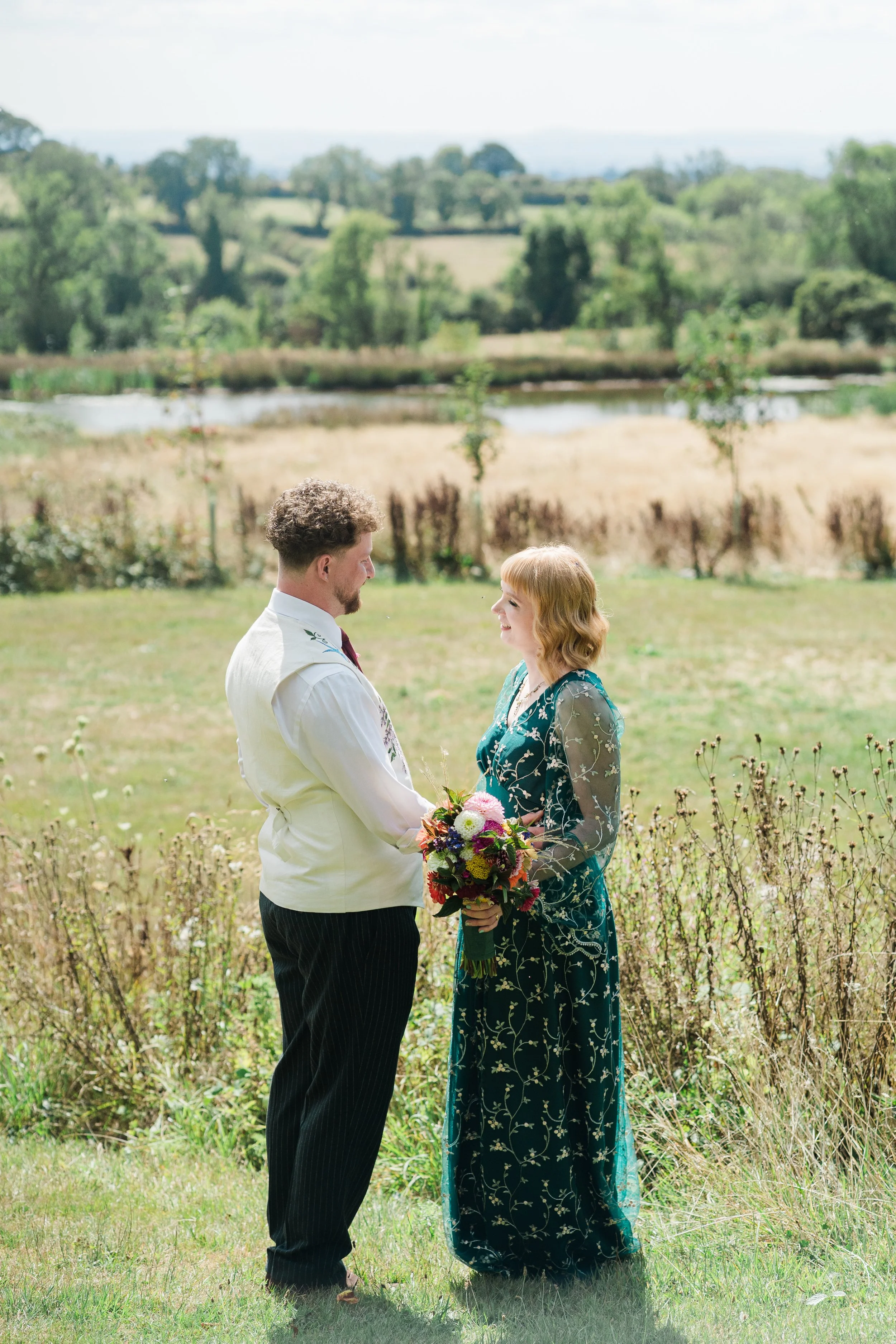 A man and woman stand facing each other outdoors in a field, holding hands with a bouquet of flowers between them, with green trees and hills in the background on a sunny day at Lapwing Farm