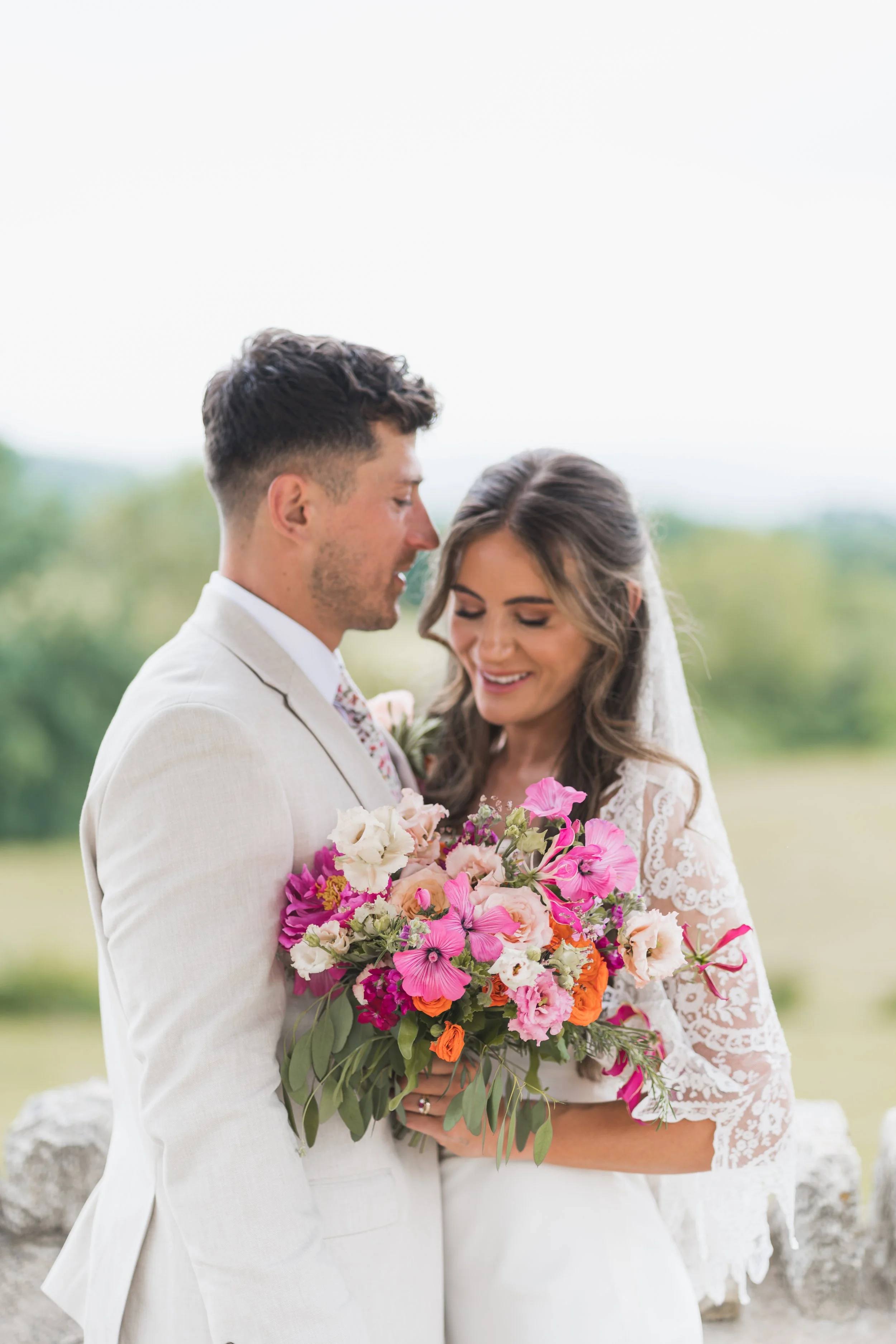 A bride and groom stand close together outdoors, holding a large bouquet of colorful flowers including pink, purple, white, and orange blooms, with blurred greenery in the background.