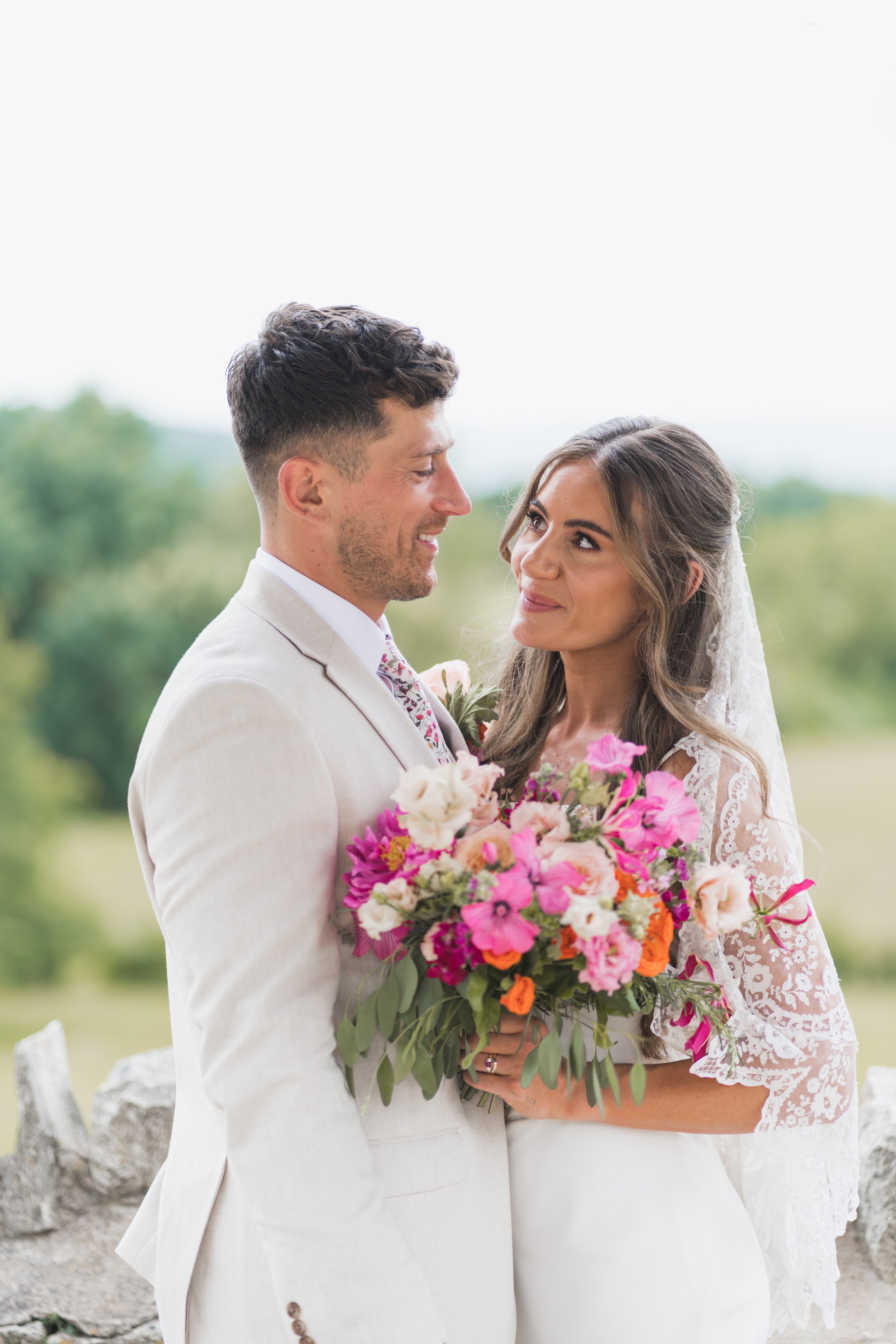 A bride and groom standing outdoors, holding a large colorful bouquet of flowers, smiling and looking at each other, with greenery in the background.