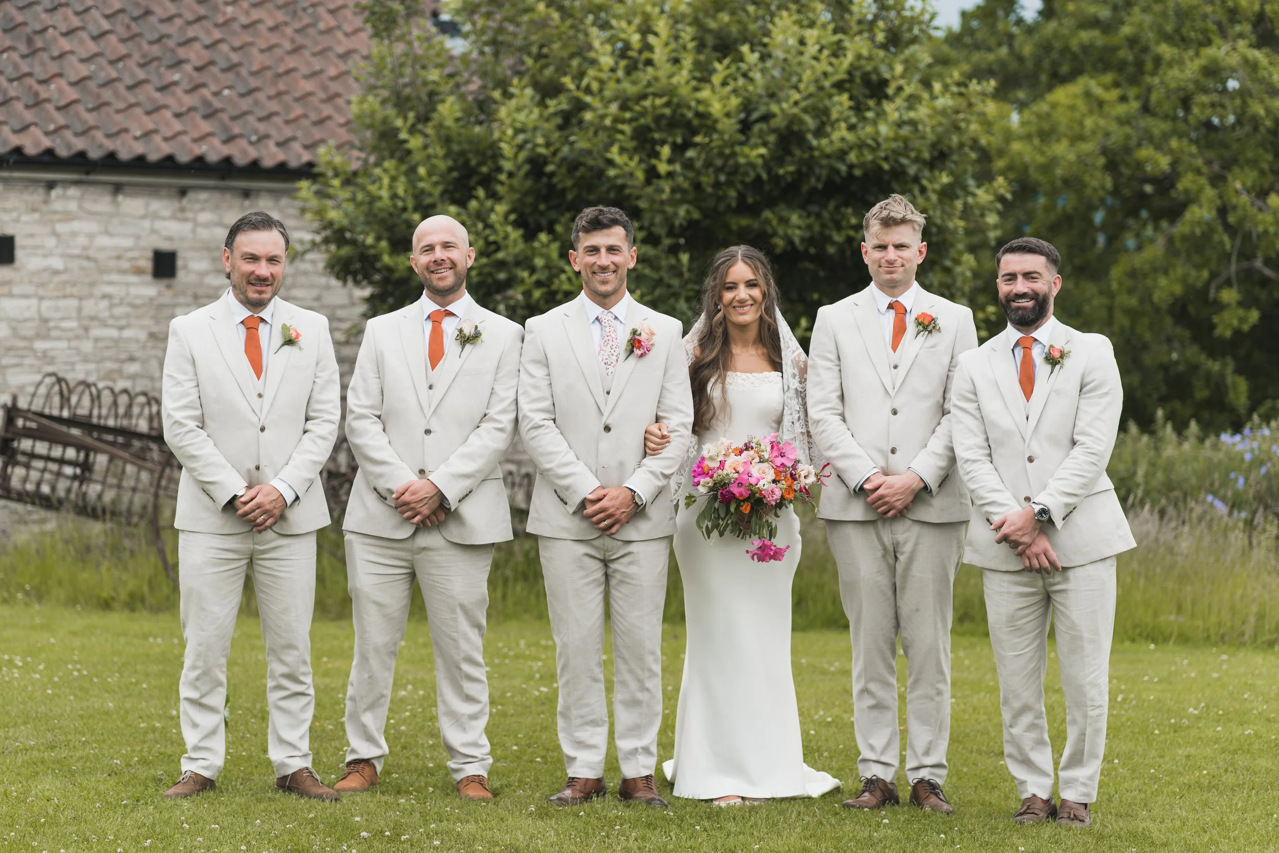 A wedding group photo of seven people standing outdoors on grass, with a rustic stone building and trees in the background. The bride, center, wears a white wedding dress holding a colorful bouquet. Six men, in white suits with orange ties and boutonnieres, stand beside her, three on each side. They are smiling and looking at the camera.