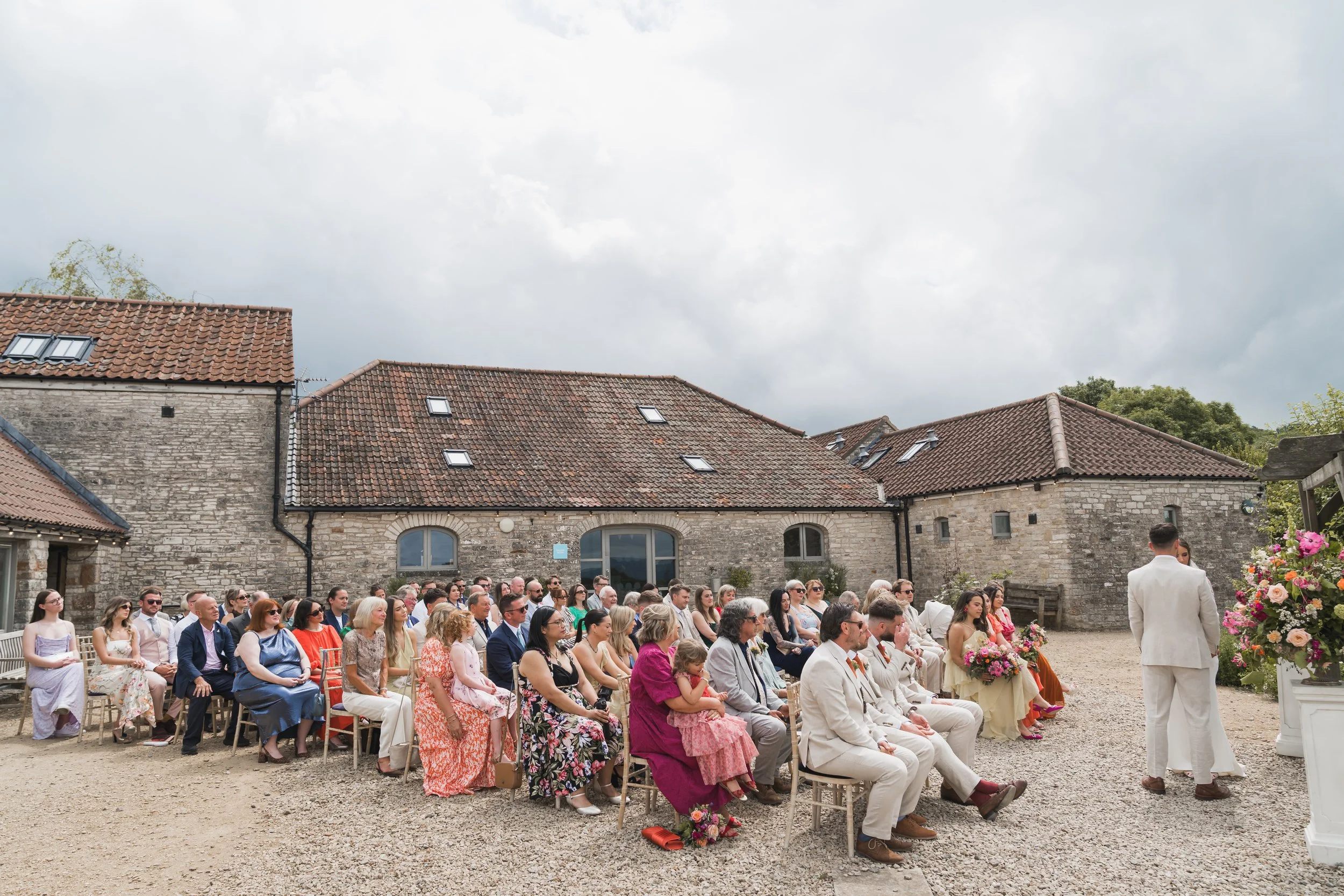 People attending a wedding ceremony outdoors in front of rustic stone buildings at Folly Farm Bristol with tiled roofs, under a cloudy sky.