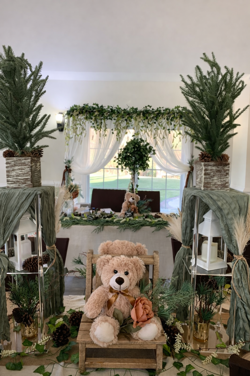 Decorated indoor space with two large potted evergreen trees, green and white draped curtains, a small plush teddy bear with a ribbon and a flower, and various greenery and pinecone accents.