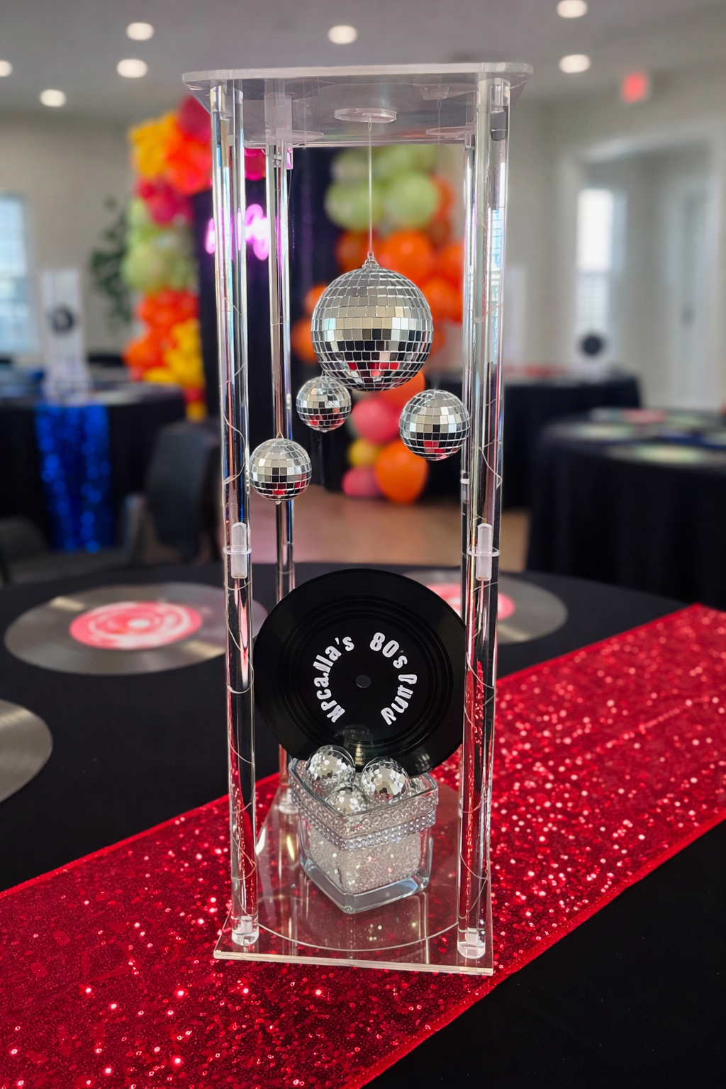Decorative centerpiece with four disco balls of varying sizes hanging inside a clear acrylic display case, set on a red glitter table runner at a celebration or party.