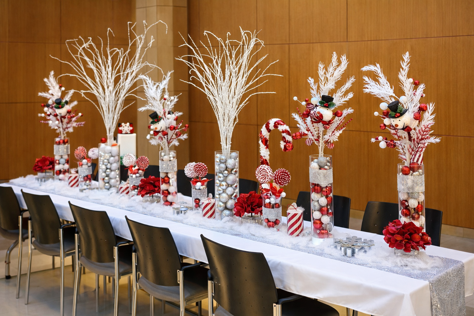 Christmas holiday table centerpiece with white branches, red ornaments, poinsettias, candy canes, and snowflake decorations on a long white table with black chairs in a decorated room.