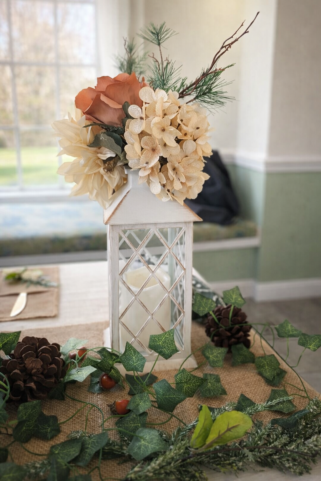 Decorative flower arrangement in a white lantern with dried white and brown flowers, pine branches, and decorative greenery, placed on a table with pinecones, ivy, and other foliage.