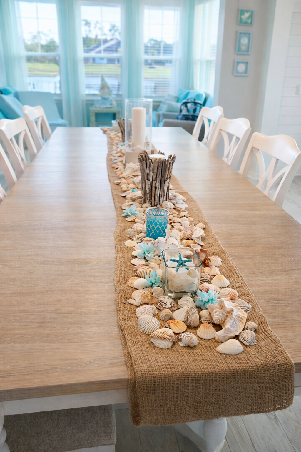 A seaside-themed dining table decorated with seashells, starfish, candles, and blue accents, with a window view of the water and a bright, airy room.