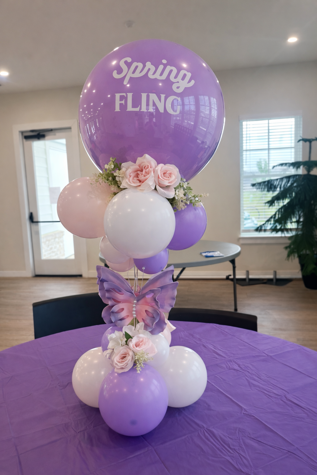 A decorative balloon arrangement with a large purple balloon that says 'Spring Fling,' surrounded by smaller pink and white balloons, pink roses, a purple butterfly-shaped ornament, and pink roses with greenery at the base. The arrangement sits on a purple tablecloth in a room with a door, window, and indoor plant in the background.