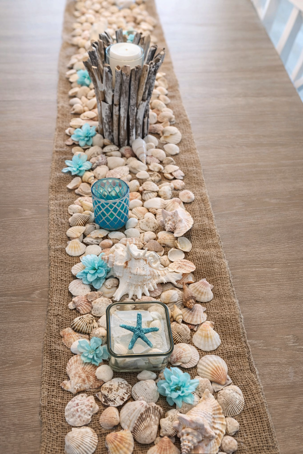 A beach-themed table runner decorated with seashells, starfish, blue flowers, candles, and driftwood centerpiece on a wooden table.