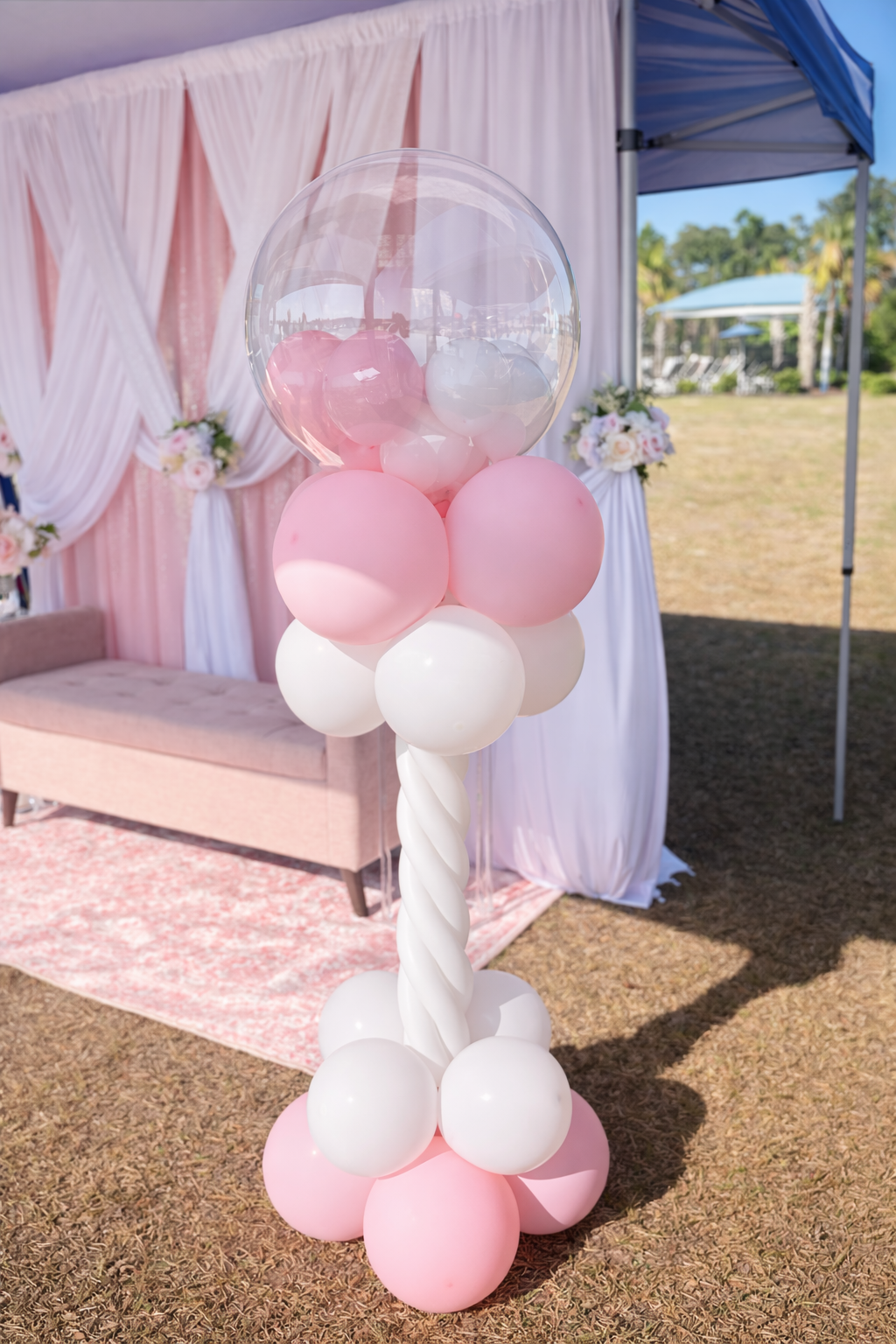Pink, white, and clear balloons arranged in a cluster, with a pink backdrop and floral decorations in the background, at an outdoor event or celebration.