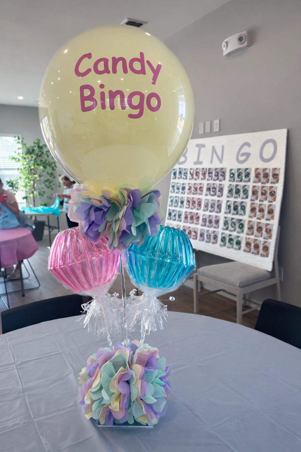Balloon centerpiece with a large yellow balloon labeled 'Candy Bingo', surrounded by pink and blue wrapped candies with tissue paper pom-poms, on a white tablecloth at a party or event.