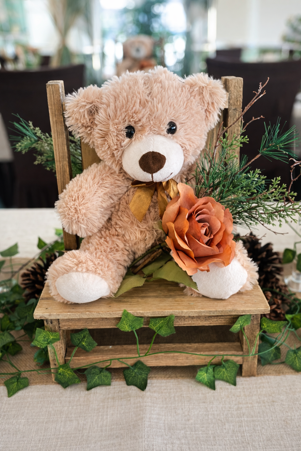A plush teddy bear with a brown nose, dark eyes, and a gold bow around its neck, sitting on a small wooden chair decorated with green foliage, pinecones, and a large orange rose, in an indoor setting with blurred background.