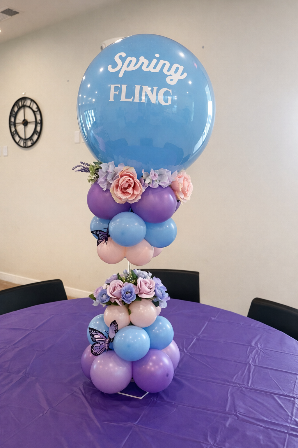 Balloon centerpiece with a large blue balloon that says 'Spring Fling', smaller purple, pink, and blue balloons, artificial flowers in pastel colors, and decorative butterfly accents, all arranged on a purple tablecloth.