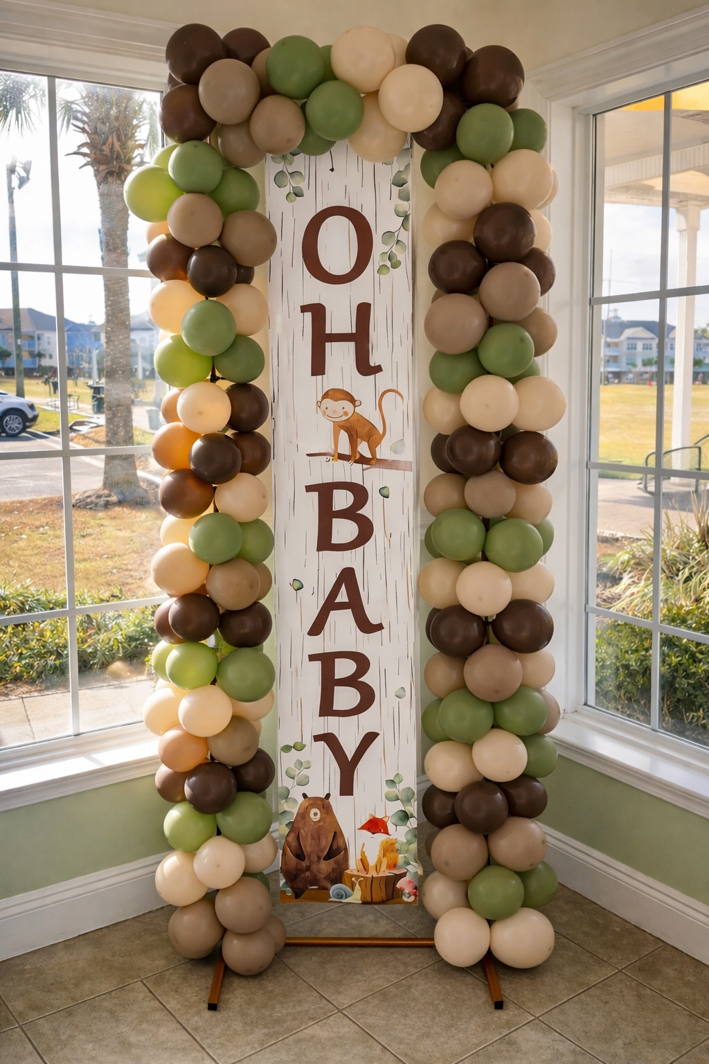 Balloon arch framing a vertical boho-themed baby shower sign with a monkey illustration, displaying the words 'Oh Baby' in brown letters, set near windows with a view of cars and houses outside.