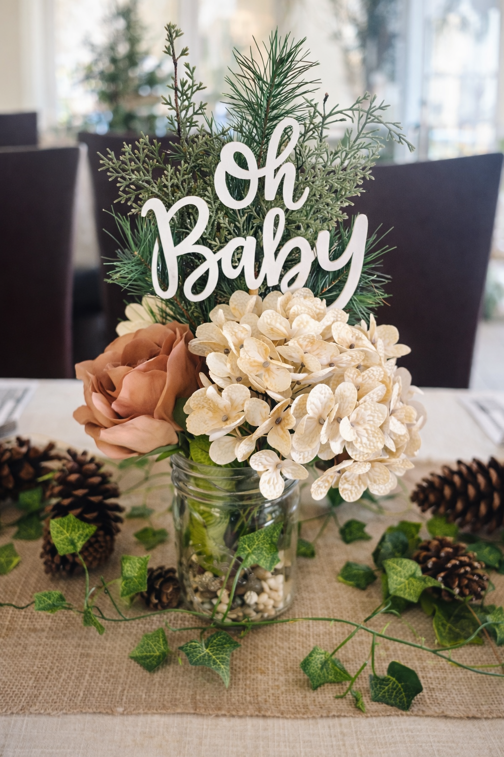 A floral arrangement in a glass jar with cream-colored hydrangeas, peach roses, green pine and ivy, and a white "Oh Baby" sign on top. The table has pinecones and ivy scattered on a burlap table runner.