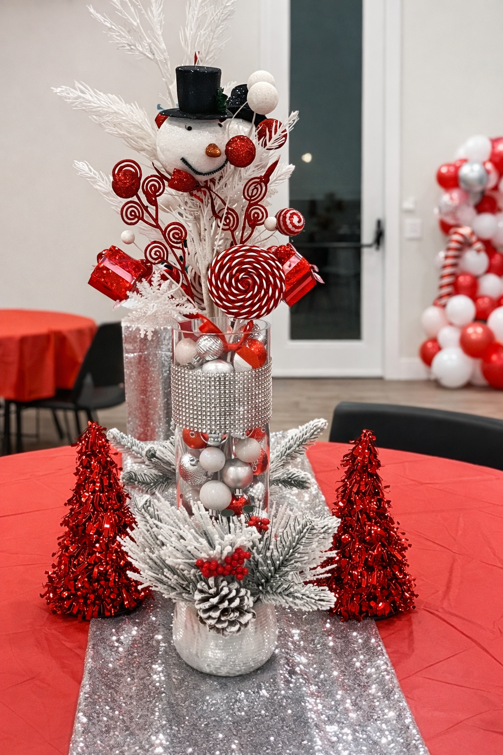 Christmas centerpiece with a snowman decoration, red and white ornaments, candy canes, red gift boxes, white pine branches, pine cones, and red metallic trees on a table with a red tablecloth and a silver glitter table runner.