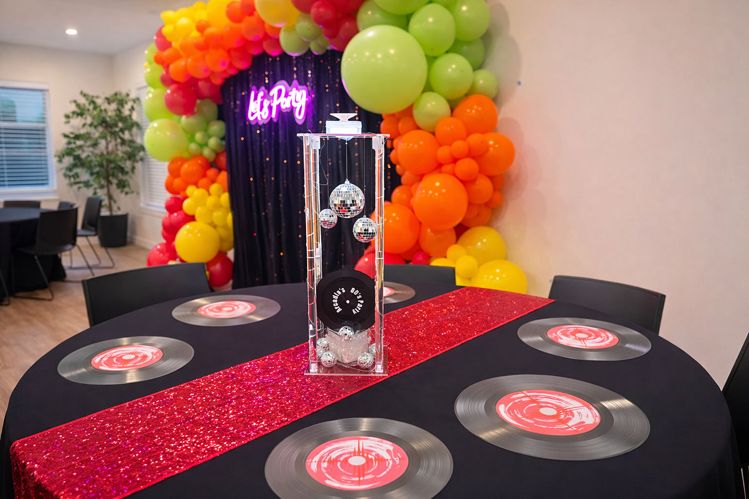 A decorated party table with a black tablecloth, circular glow-in-the-dark centerpieces, and a red glittery table runner. A central decor piece features disco balls and a weight labeled "80S PARTY". Behind the table is a colorful balloon arch with shades of green, yellow, orange, and red, framing a backdrop with neon "Let's Party" sign. The room has wood flooring, a window, and a potted plant.
