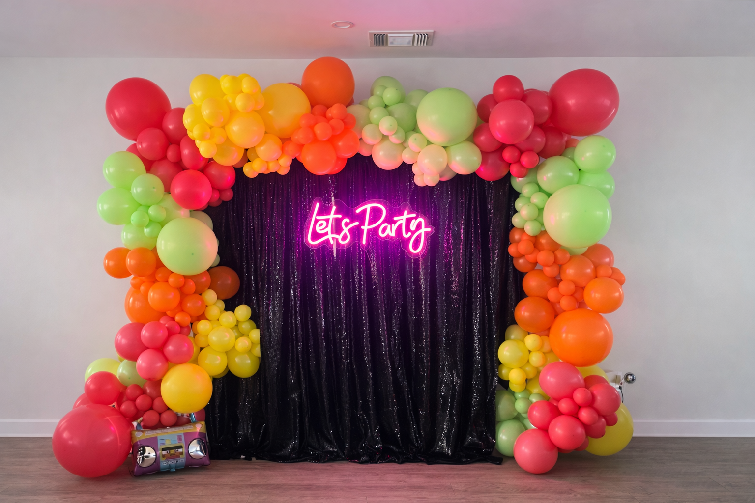 Colorful balloon arch with pink, yellow, orange, and green balloons framing a dark purple curtain backdrop with a neon pink sign that reads 'Let's Party'.