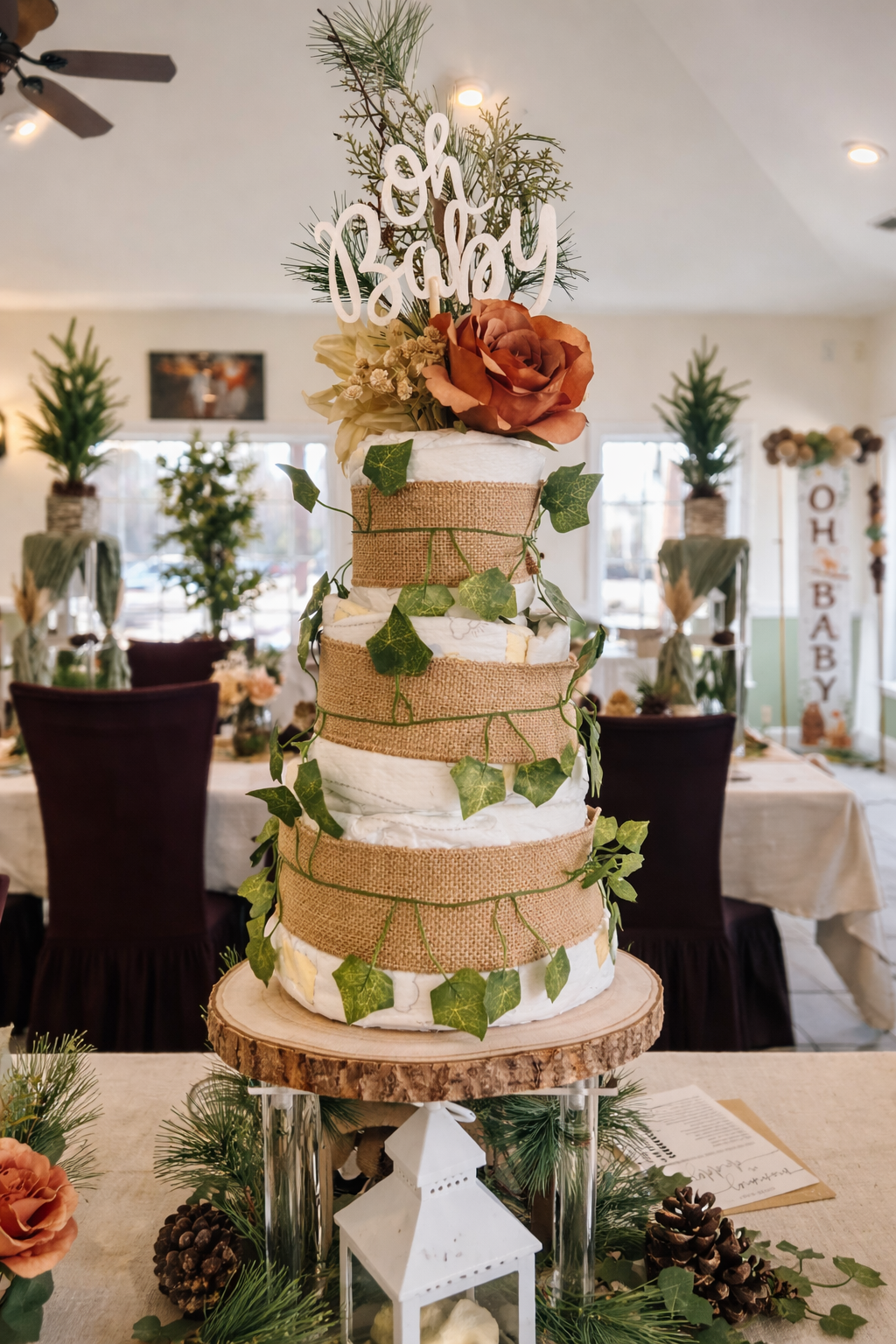 A tiered wedding cake decorated with rustic burlap ribbon and green ivy leaves, topped with a floral arrangement and a 'Oh Baby' cake topper, set on a wooden slab stand inside a decorated reception hall.