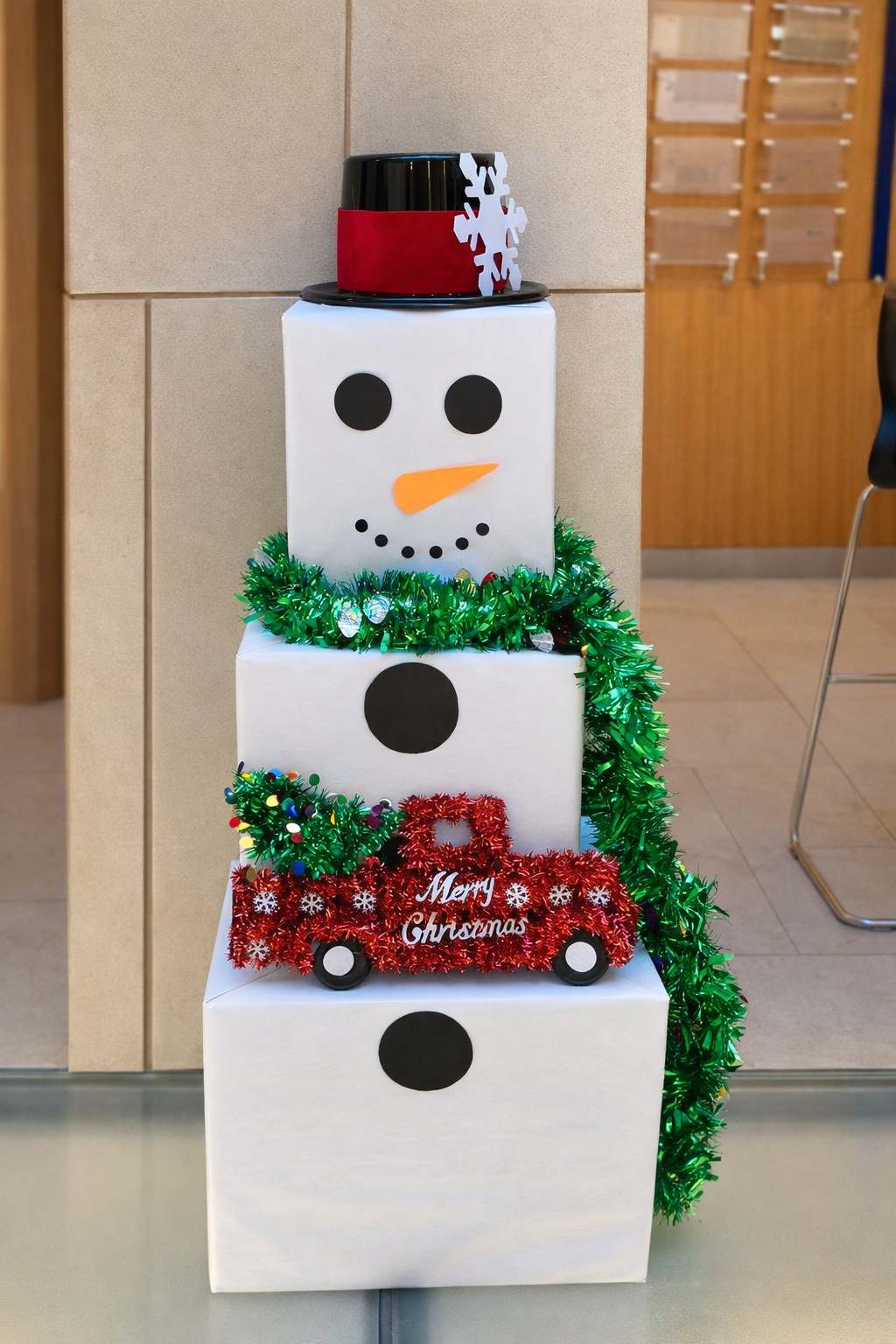 Decorative stack of four white boxes arranged to resemble a snowman's face, with black circle eyes, an orange carrot nose, and a black dotted smile, adorned with festive Christmas decorations including a green tinsel wreath, red tinsel wagon with 'Merry Christmas' sign, and topped with a black hat decorated with a red band and a white snowflake, set against a neutral background.