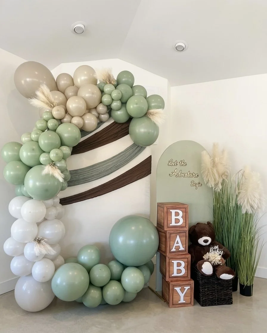 Decorative baby shower setup with green, white, and beige balloons arranged in a large, cascading arch, teddy bear, wooden blocks spelling 'BABY', and tall pampas grass in black pots.