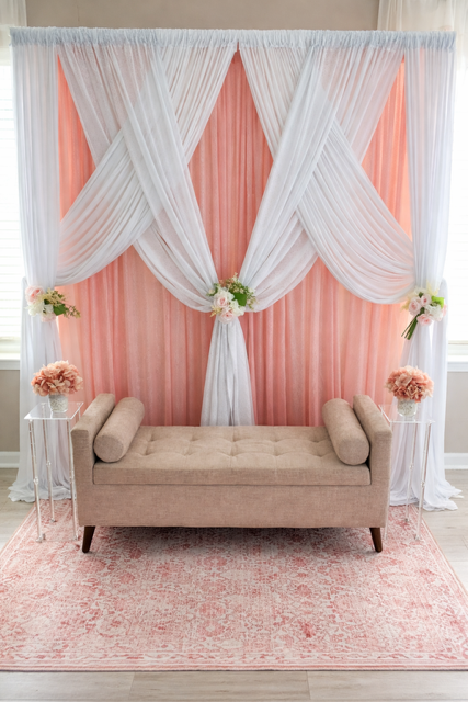 A beige sofa in front of a pink and white draped curtain backdrop, with side tables holding pink flower arrangements, on a pink area rug.