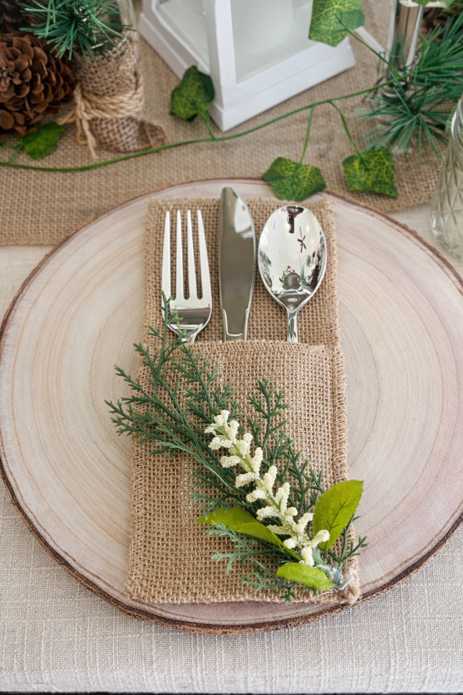 Close-up of a place setting on a round, wooden slice that acts as a plate. Includes a fork, knife, and spoon placed on a brown burlap napkin with a green leafy sprig and small white flowers. The table is decorated with greenery and a lantern in the background.