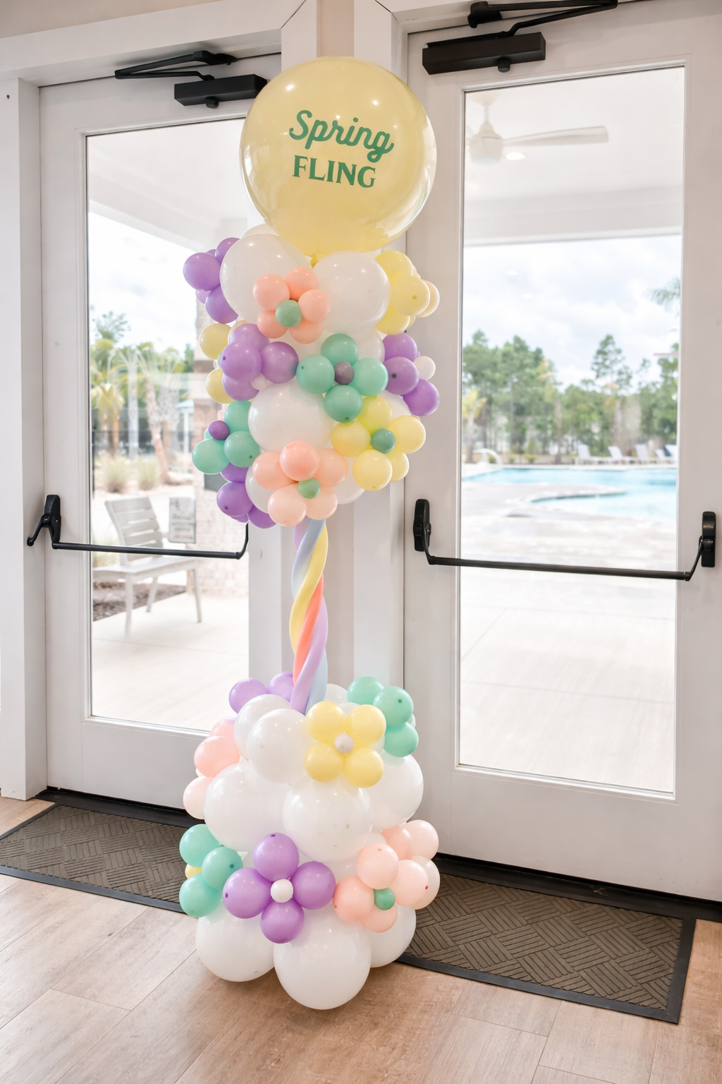 Colorful balloon arrangement with a large yellow balloon at the top reading "Spring Fling" in green, placed indoors near glass doors with a view of an outdoor pool area.