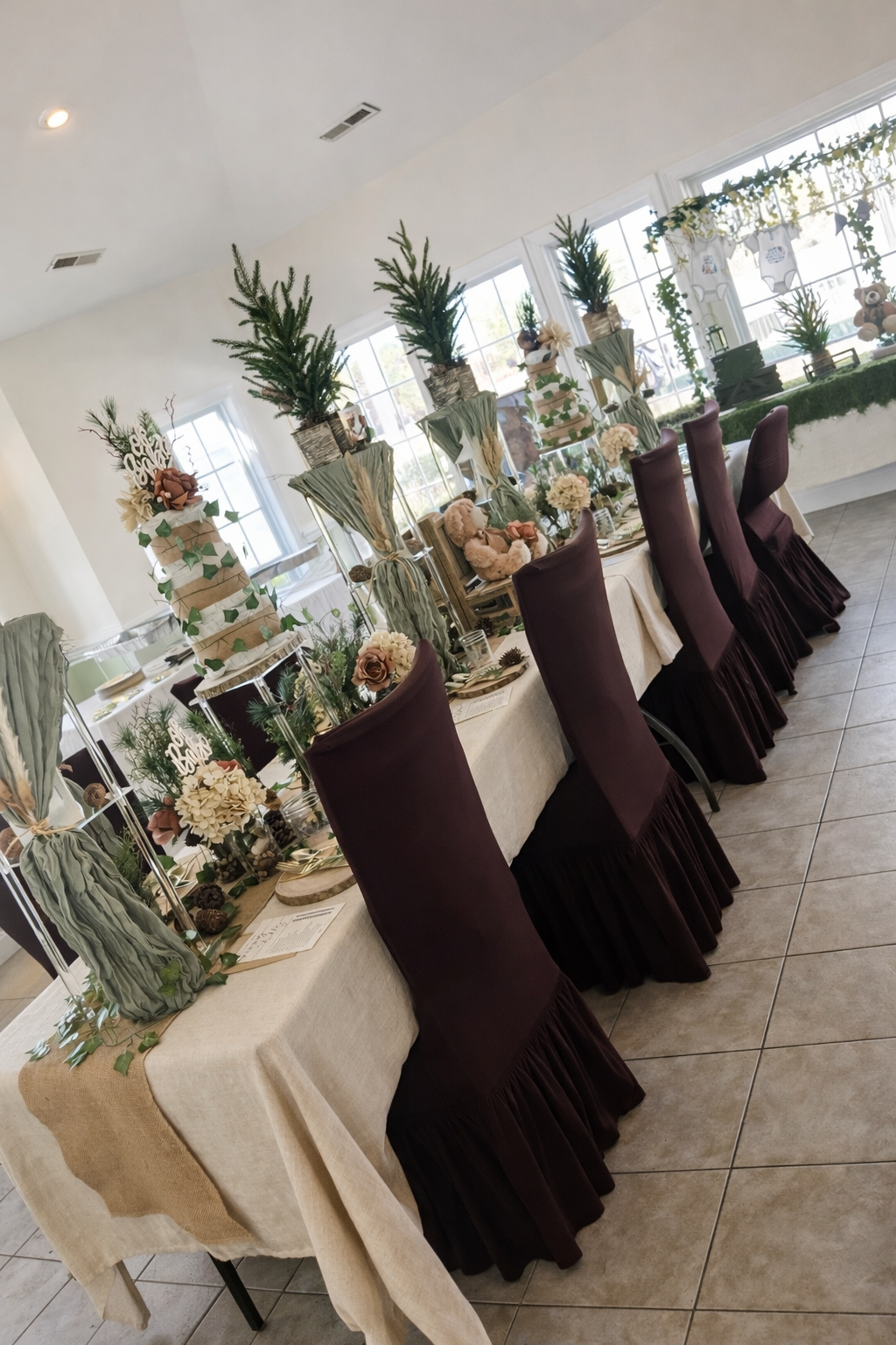 Decorated dining table with purple chairs, floral arrangements, teddy bears, and a tiered centerpiece in a bright room with large windows.