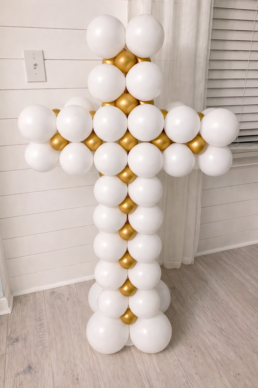 Balloon cross made of white and gold balloons positioned indoors near a white paneled wall and a window with sheer curtains.