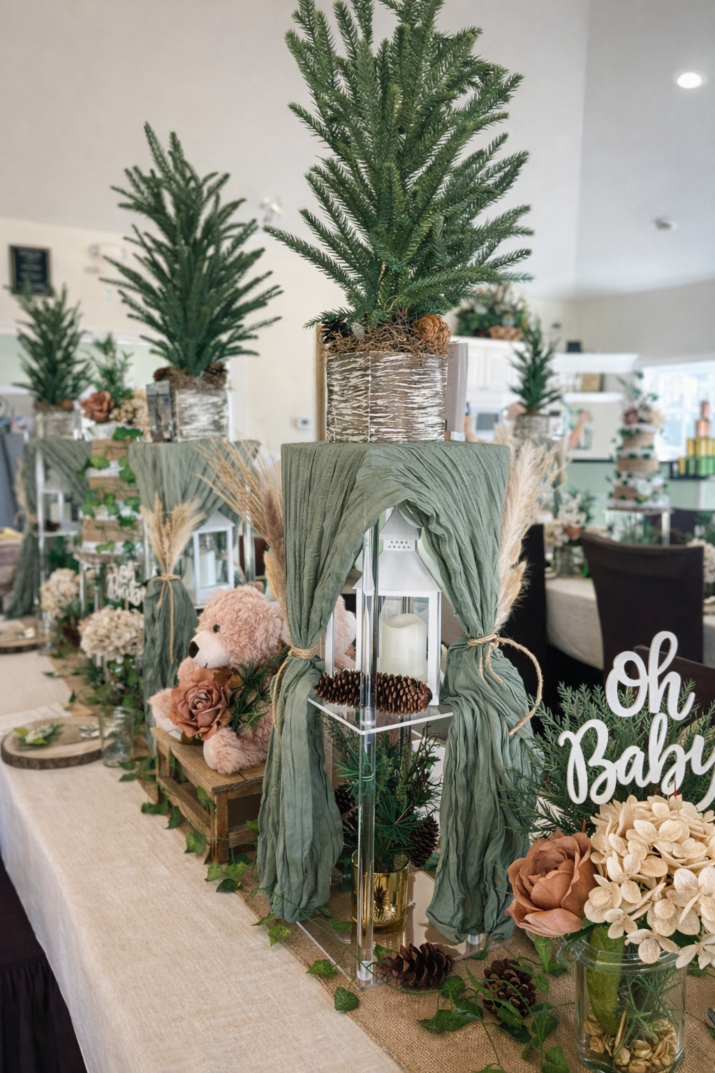 Decorative table arrangement with pine trees in vases, a teddy bear, flowers, candles, pinecones, and greenery for a baby shower celebration.