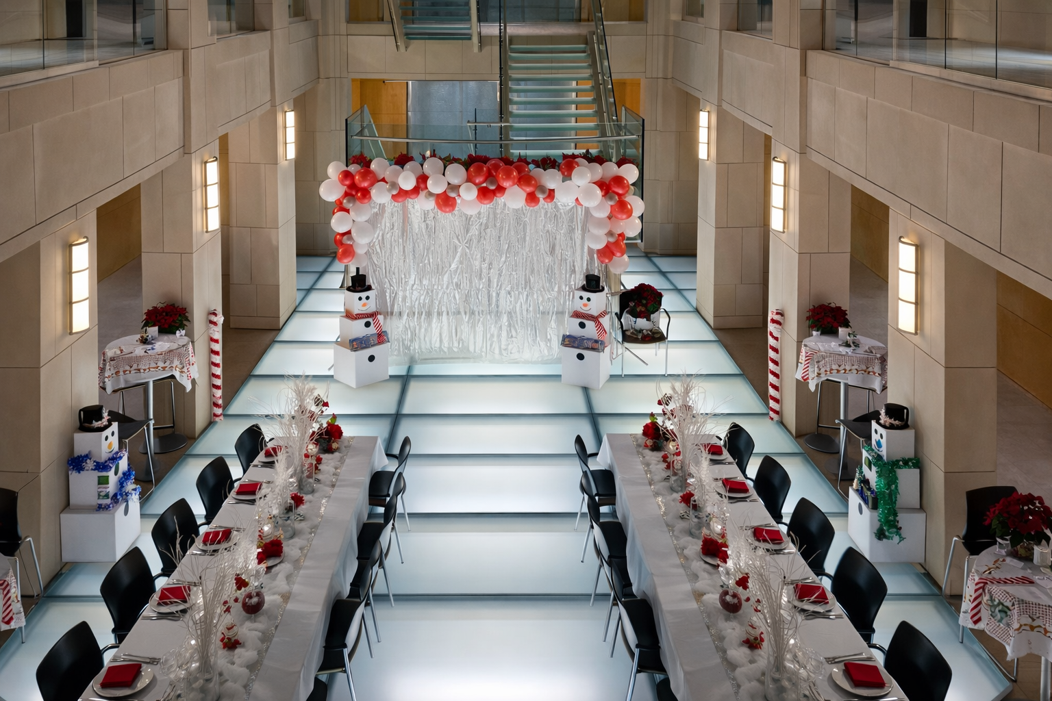 A decorated indoor space with holiday-themed decorations, including snowman figures, balloons, poinsettias, and table settings for a festive event.