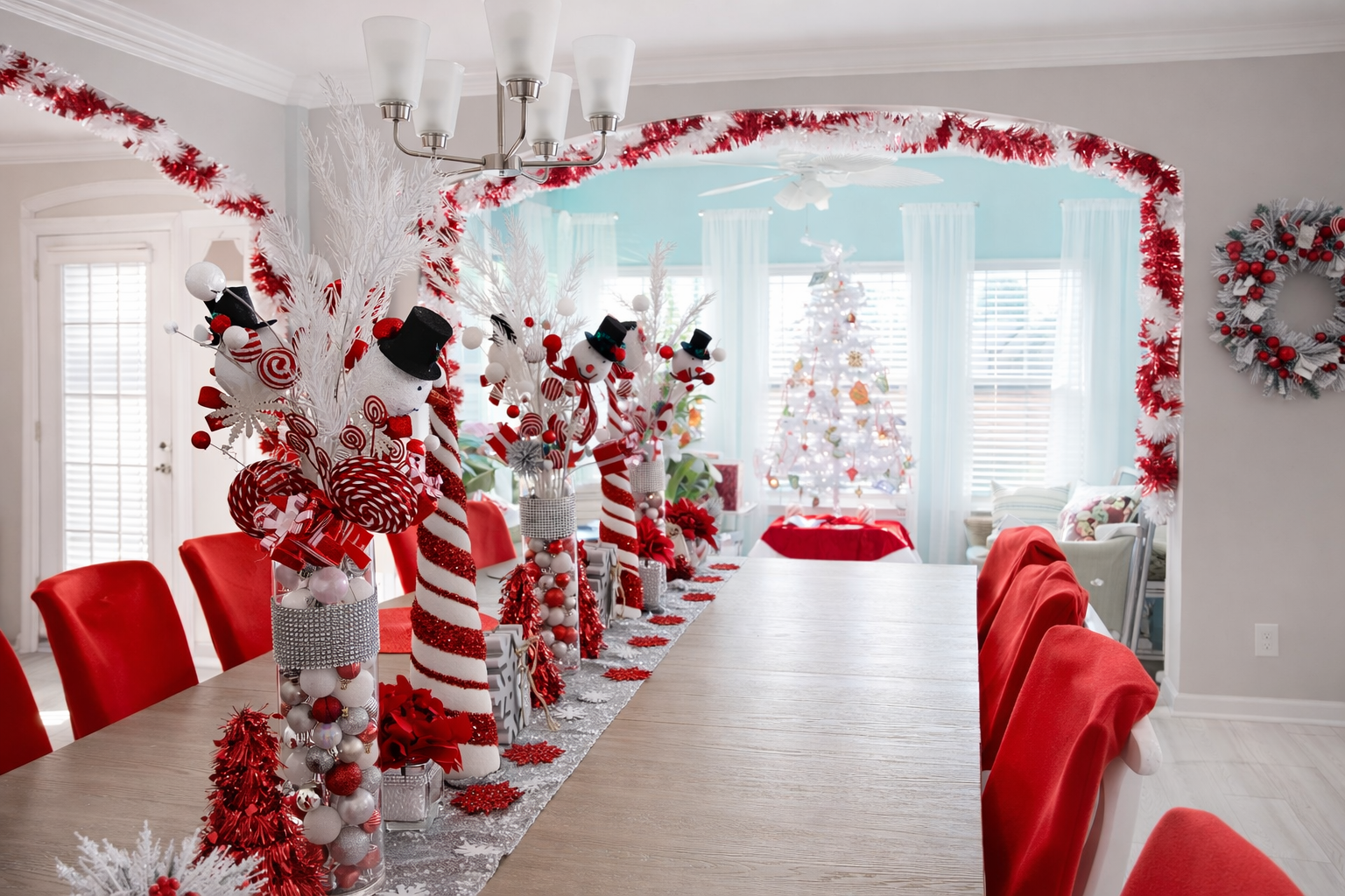 A dining room decorated for Christmas with a long table, red chair covers, and red and white holiday decorations including snowmen, candy canes, ornaments, and a Christmas tree in the background.