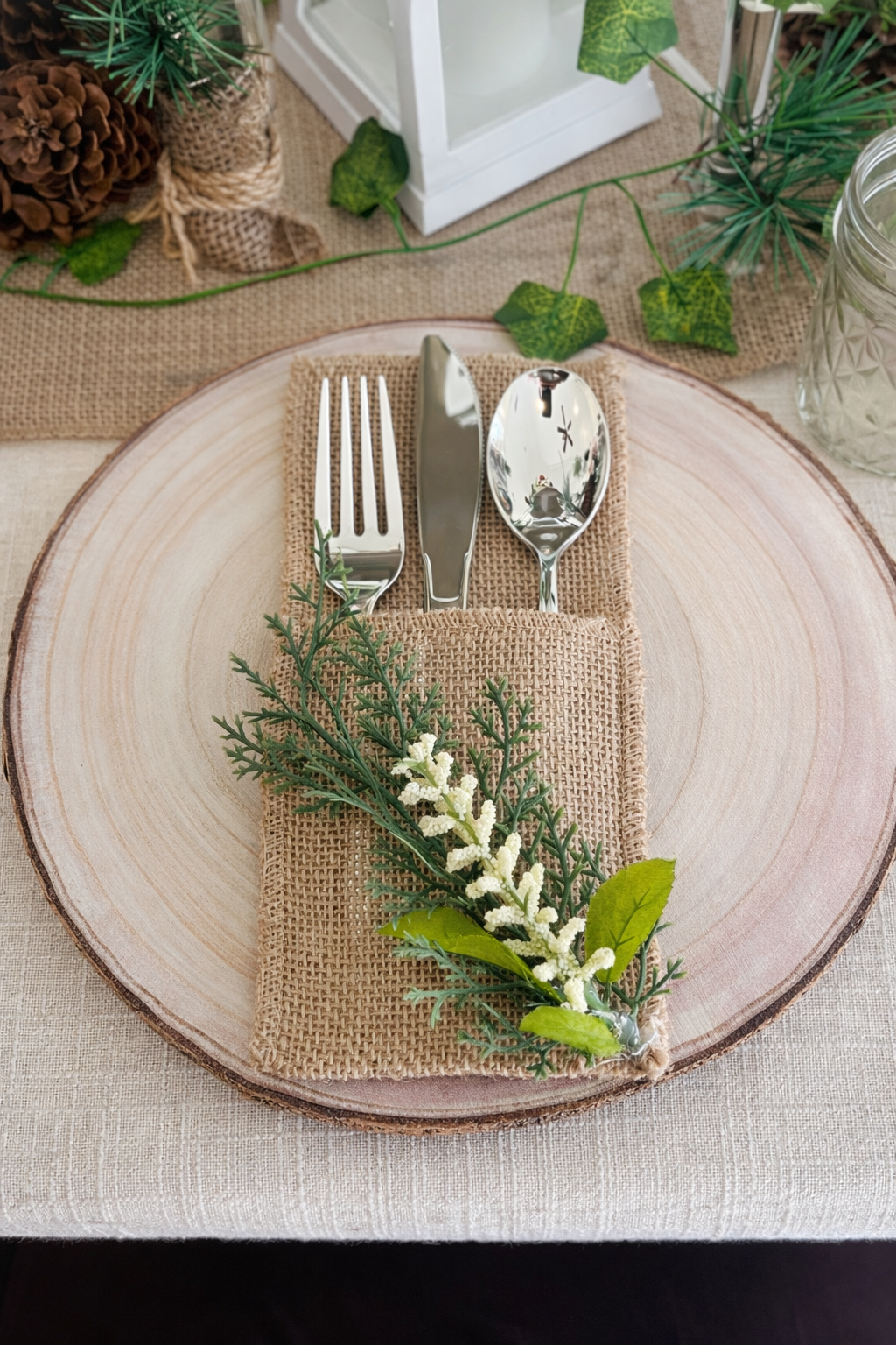 A rustic table setting with a round wooden charger plate, beige burlap napkin, and silverware consisting of a fork, knife, and spoon. The napkin is decorated with greenery and white flowers, with additional greenery and decorative items in the background.