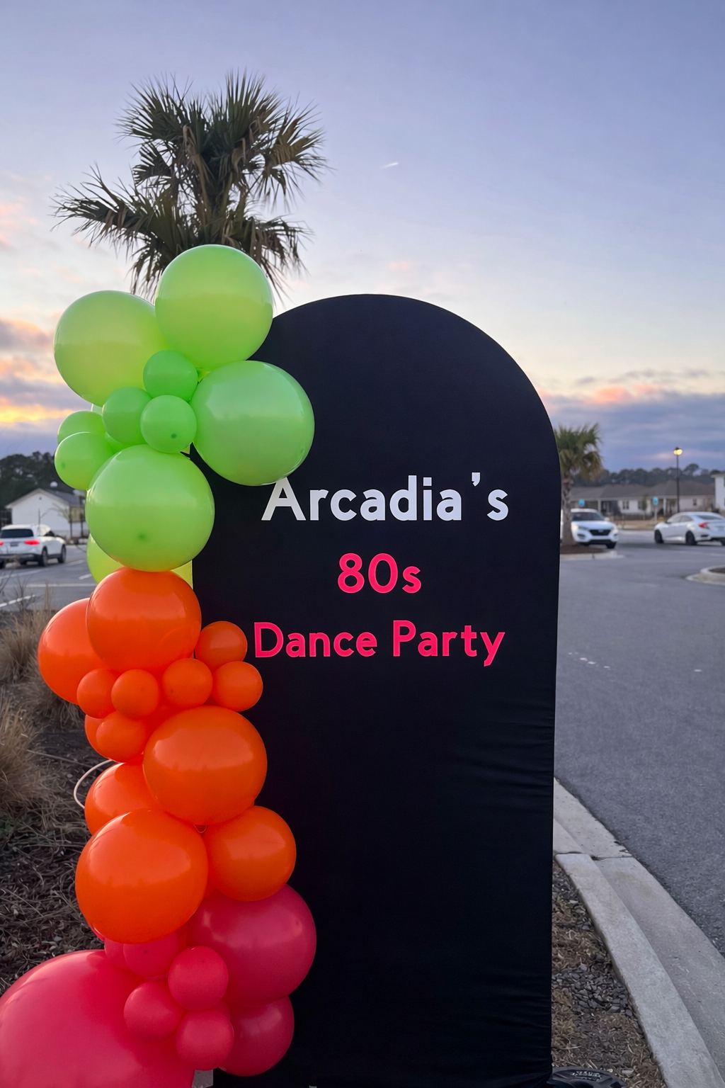 Sign for Arcadia's 80s dance party decorated with green, orange, and pink balloons, with palm trees and a parking lot in the background at sunset.