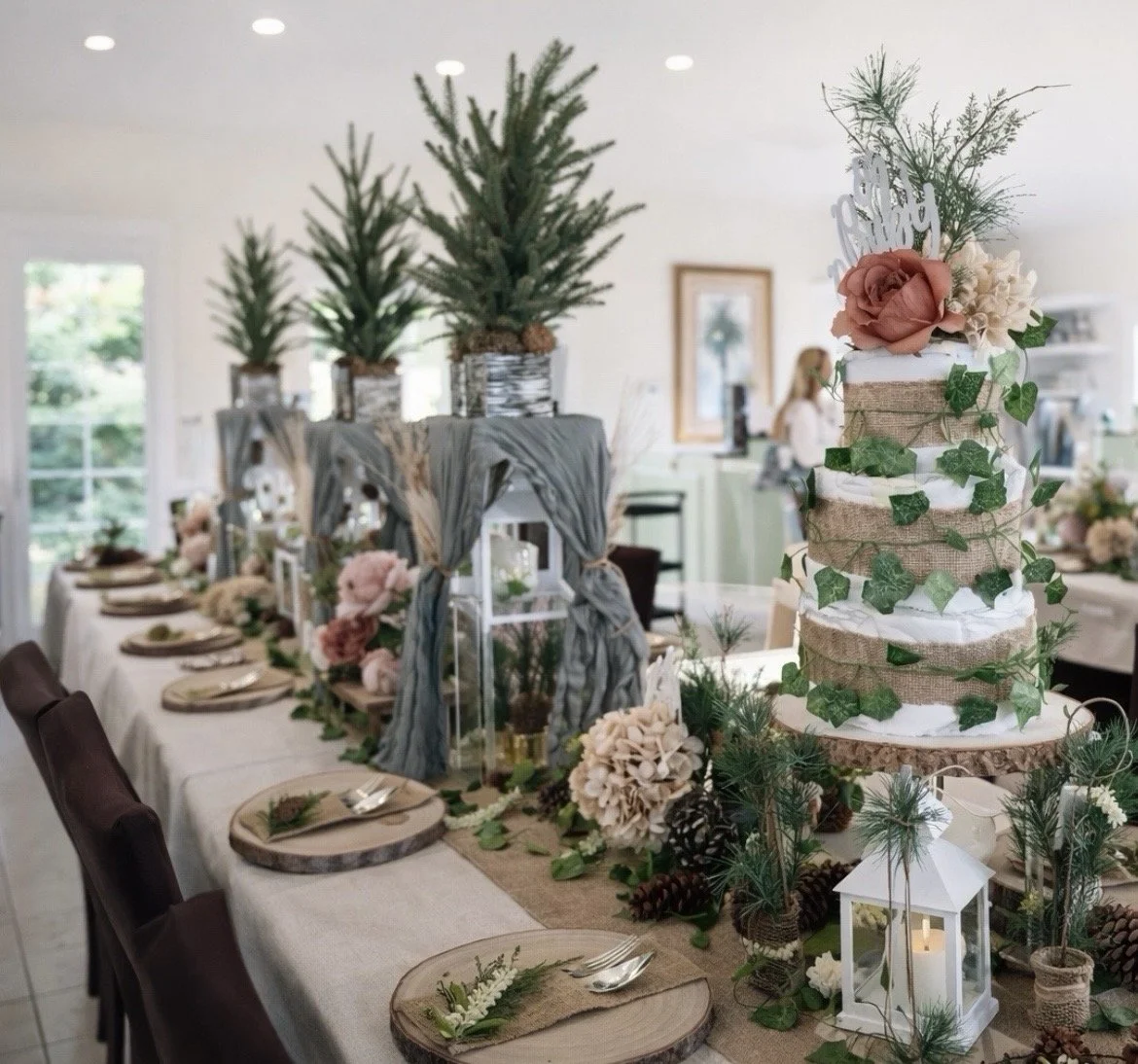 A wedding reception table decorated with a rustic theme. There is a tall, multi-layered cake decorated with green ivy, pink flowers, and a 'Happy Wedding' topper. The table has pinecones, greenery, lanterns, and flowers, with place settings and wooden charger plates.