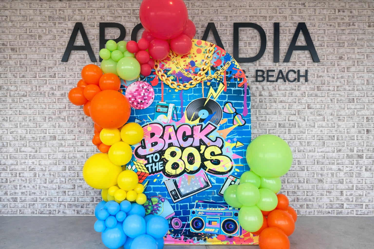 Colorful decoration for 1980s themed event featuring balloons, graffiti-style backdrop with music and pop culture icons, and the text 'Back to the 80s' on a blue brick wall.