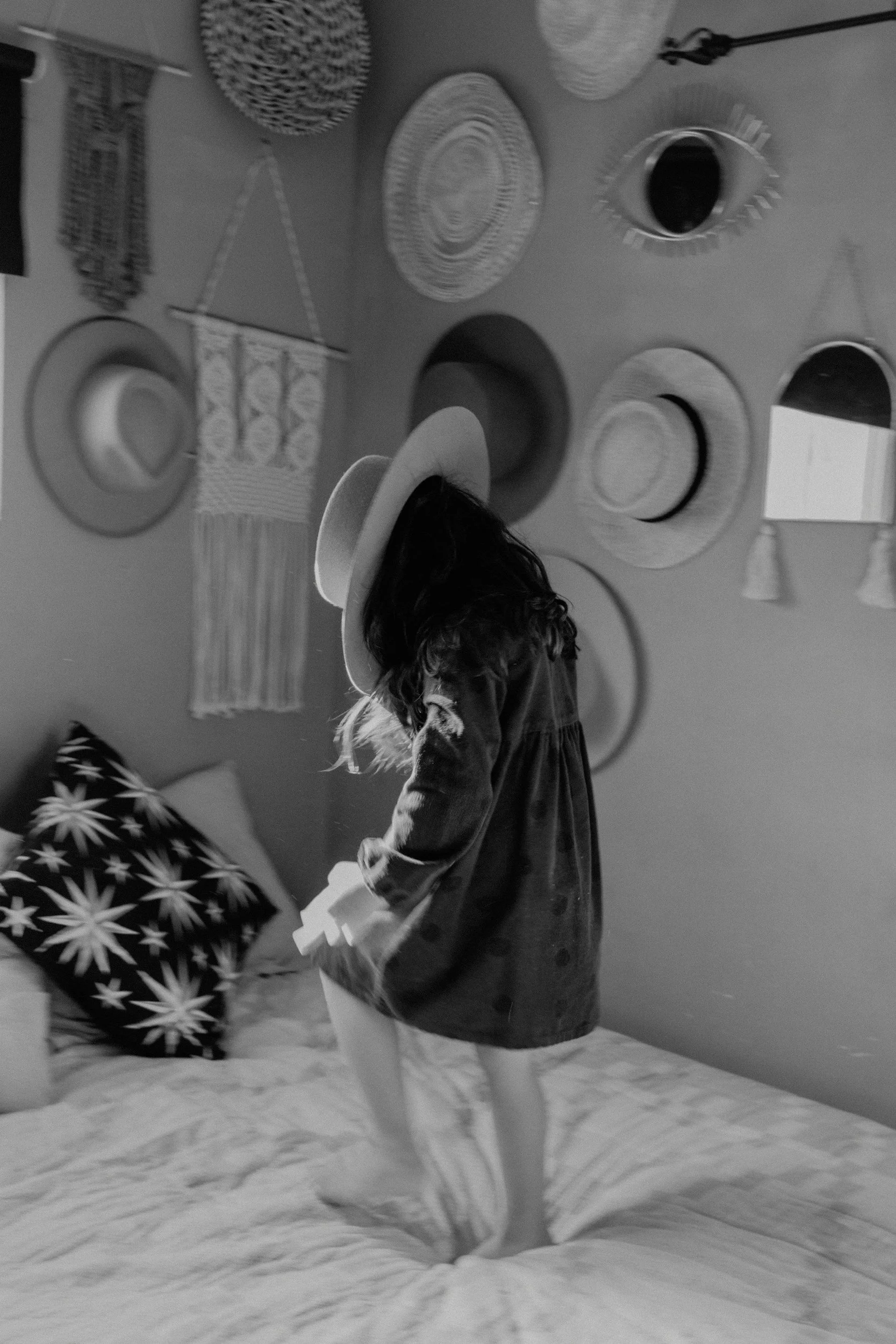 A girl wearing a wide-brimmed hat and a dress, standing on a bed with one foot, in a room decorated with woven wall hangings and hats on the wall.