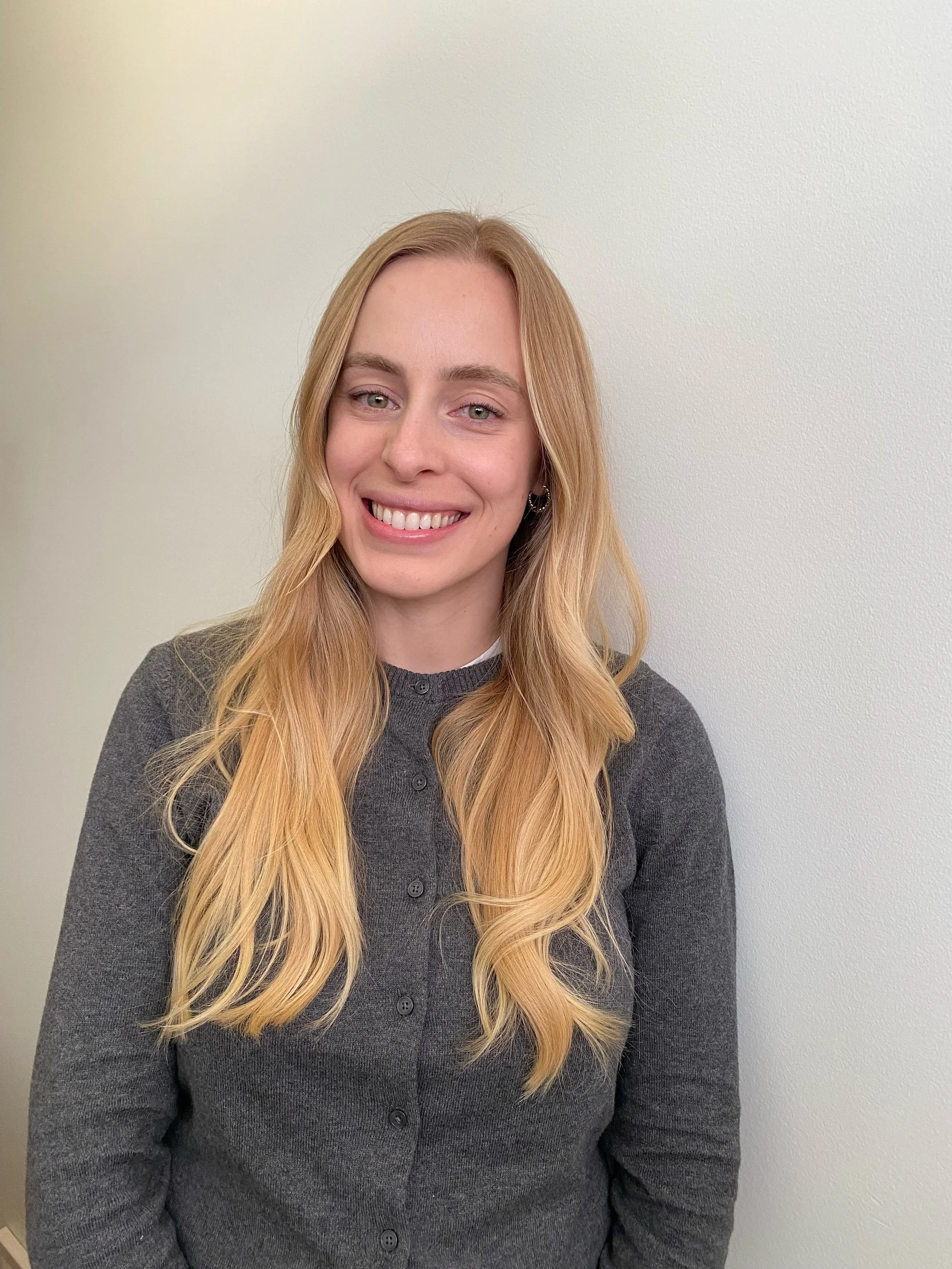 A woman with long blonde hair, lightly styled, wearing a gray cardigan, smiling, standing against a plain off-white wall.