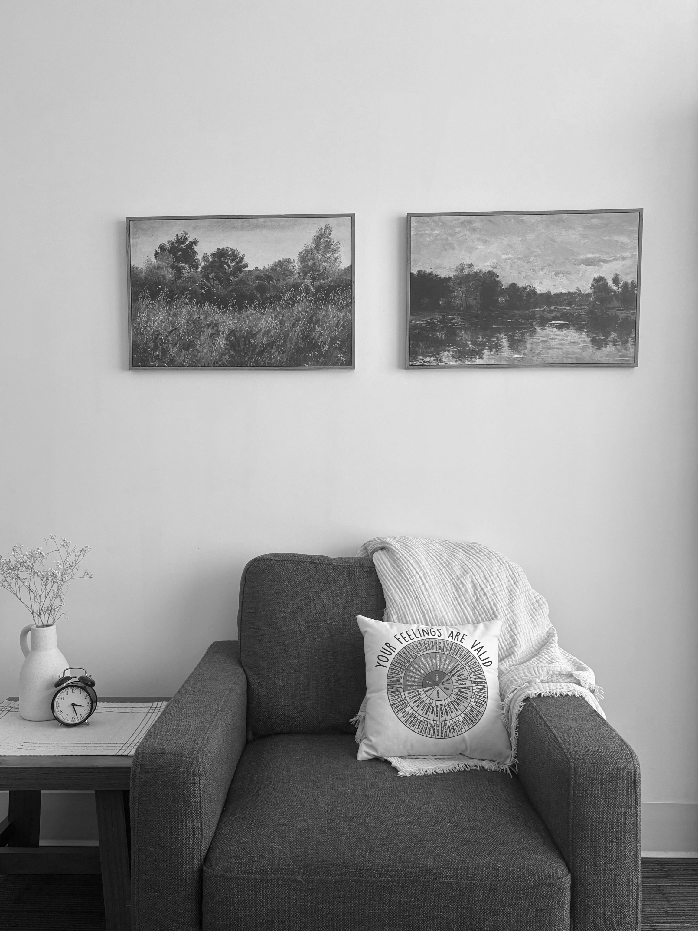 A living room with a dark gray armchair, a throw blanket, and a decorative pillow that reads 'Your feelings are valid'. Next to the armchair is a small wooden side table with a white vase containing dried flowers, a clock, and a striped table runner. Behind the armchair is a plain white wall with two landscape paintings hanging side by side.