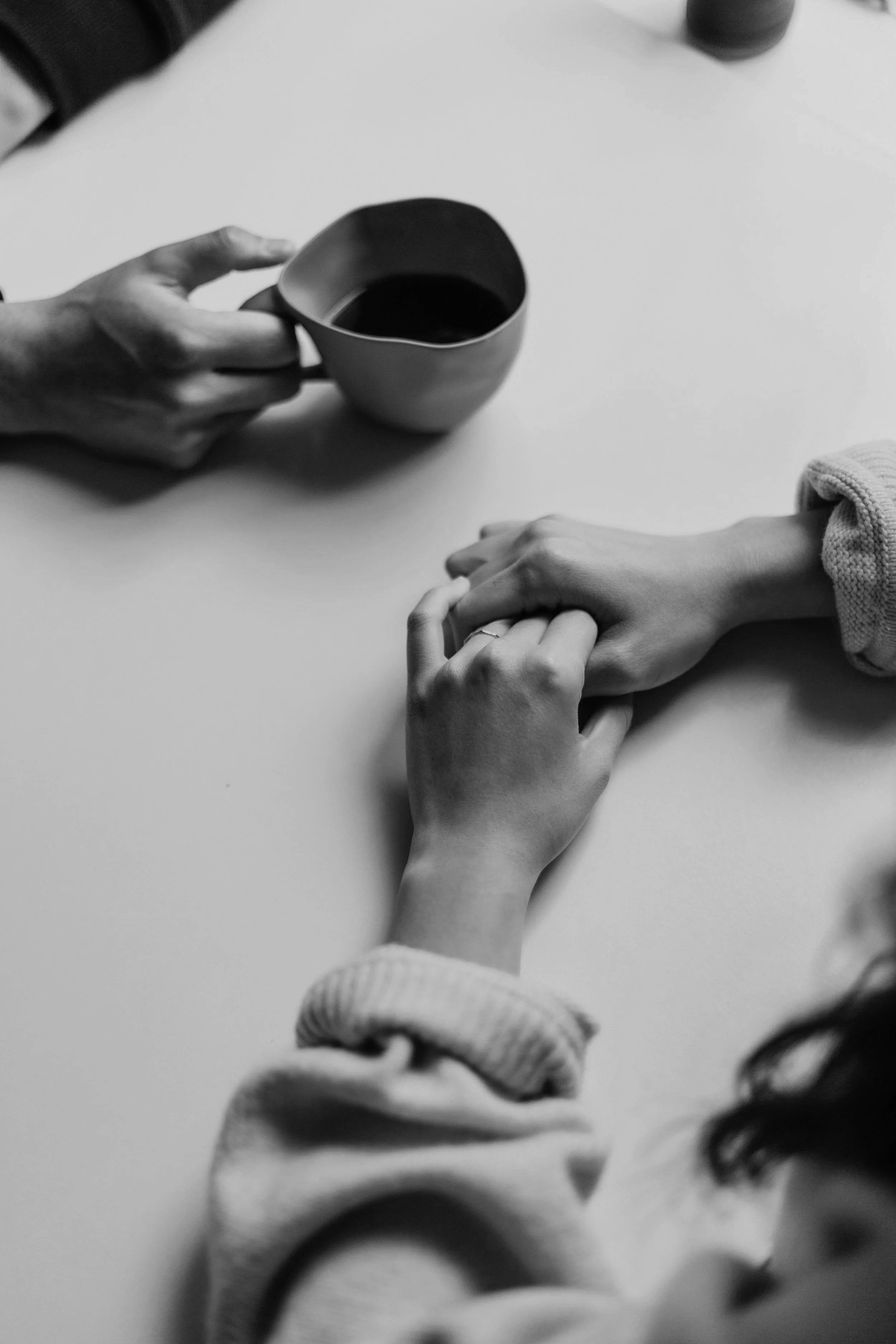 Two people holding hands across a table, with a coffee mug in the foreground. The image is in black and white.