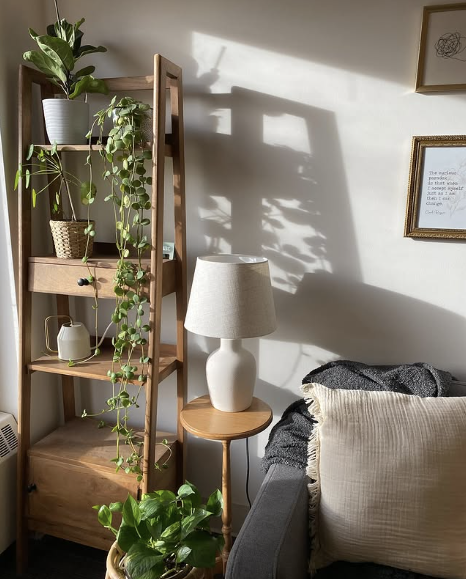 Sunlight casts shadows of houseplants on a white wall in a cozy corner of a living room with minimal decor, including a wooden shelf with potted plants, a white table lamp on a small round wooden table, and framed wall art.