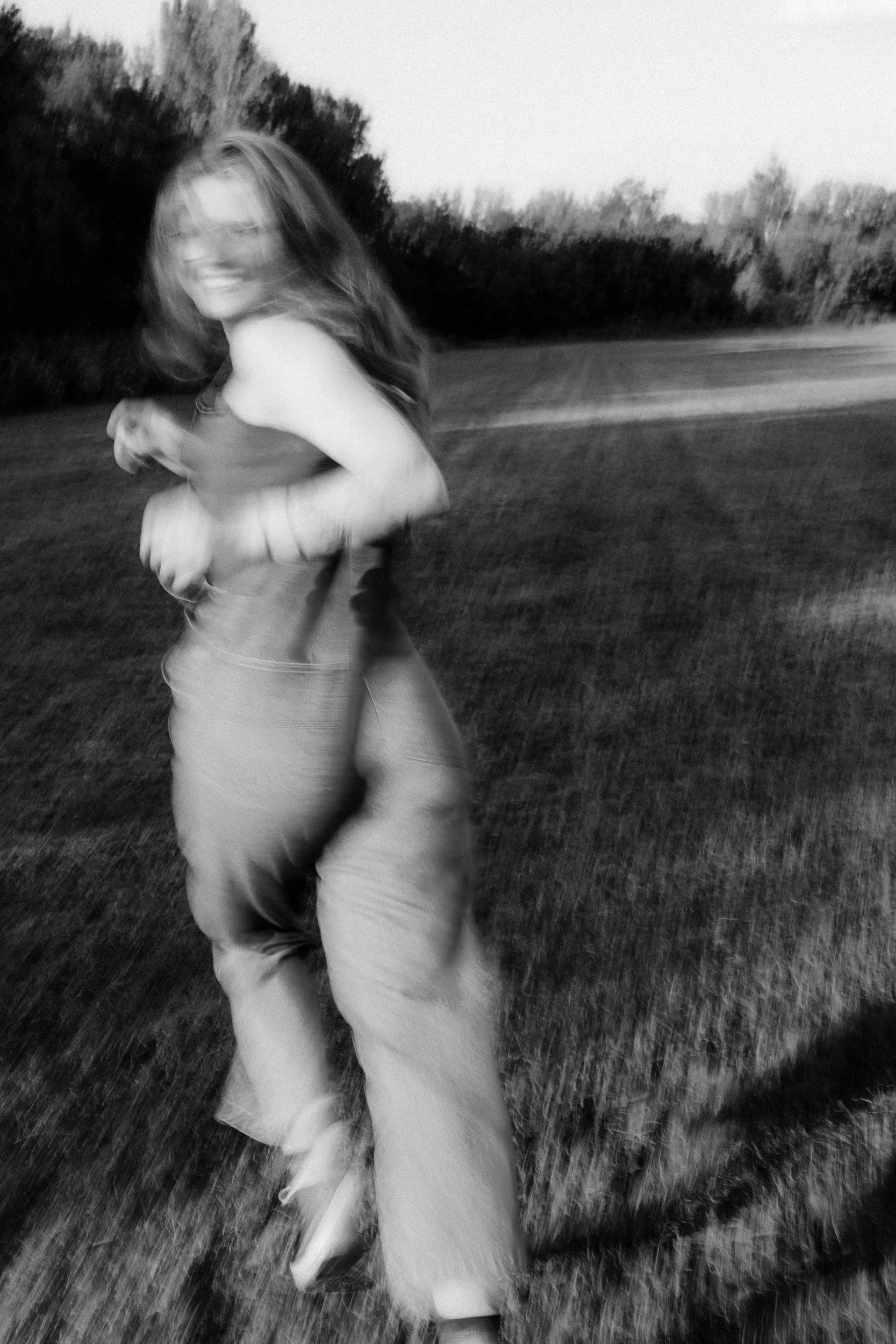 A woman with long hair running in an open grassy field during dusk or dawn, with trees in the background, captured in motion creating a blurred, artistic effect.