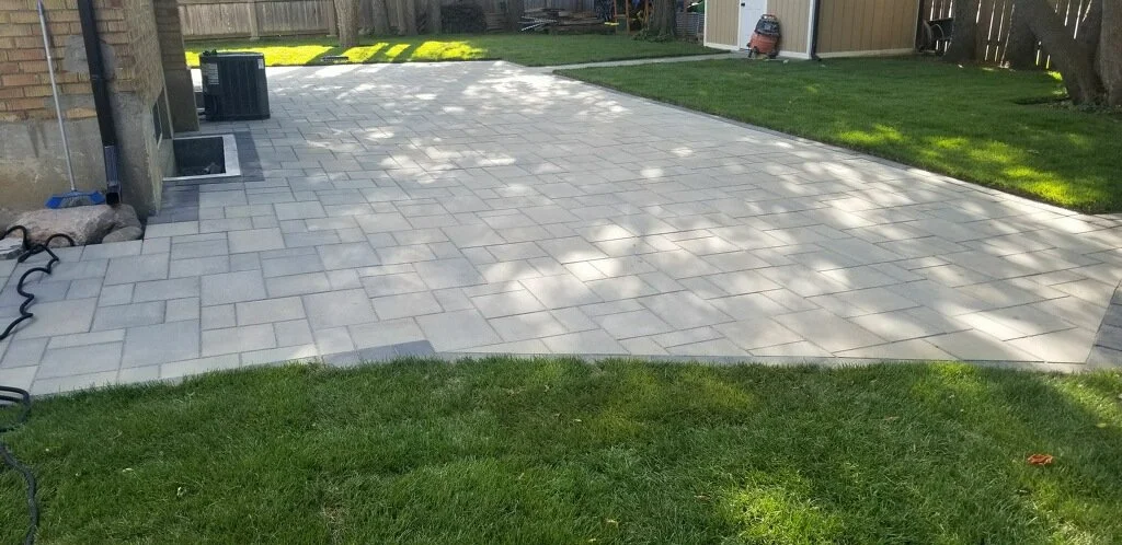 Backyard patio with light gray pavers, lush green grass in the foreground, and a wooden fence and shed in the background.