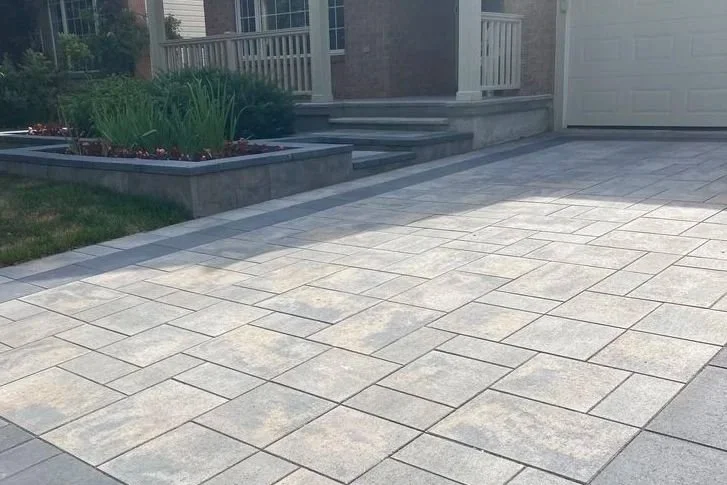 Paved driveway with light gray stone tiles next to a house with stairs, a porch with white railing, and a garden bed with plants.